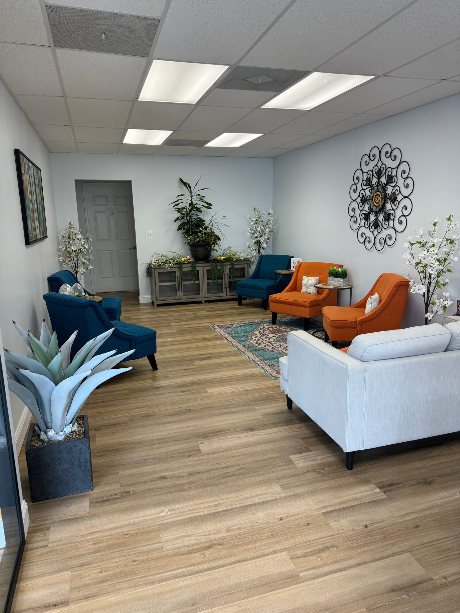 Living room with white walls, wooden floor, blue and orange furniture, plants, and decorative wall art.
