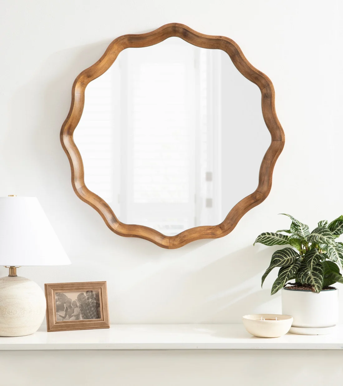 Decorative wooden mirror with a wavy frame hanging on a white wall, with a shelf below holding a lamp, picture frame, and plant.