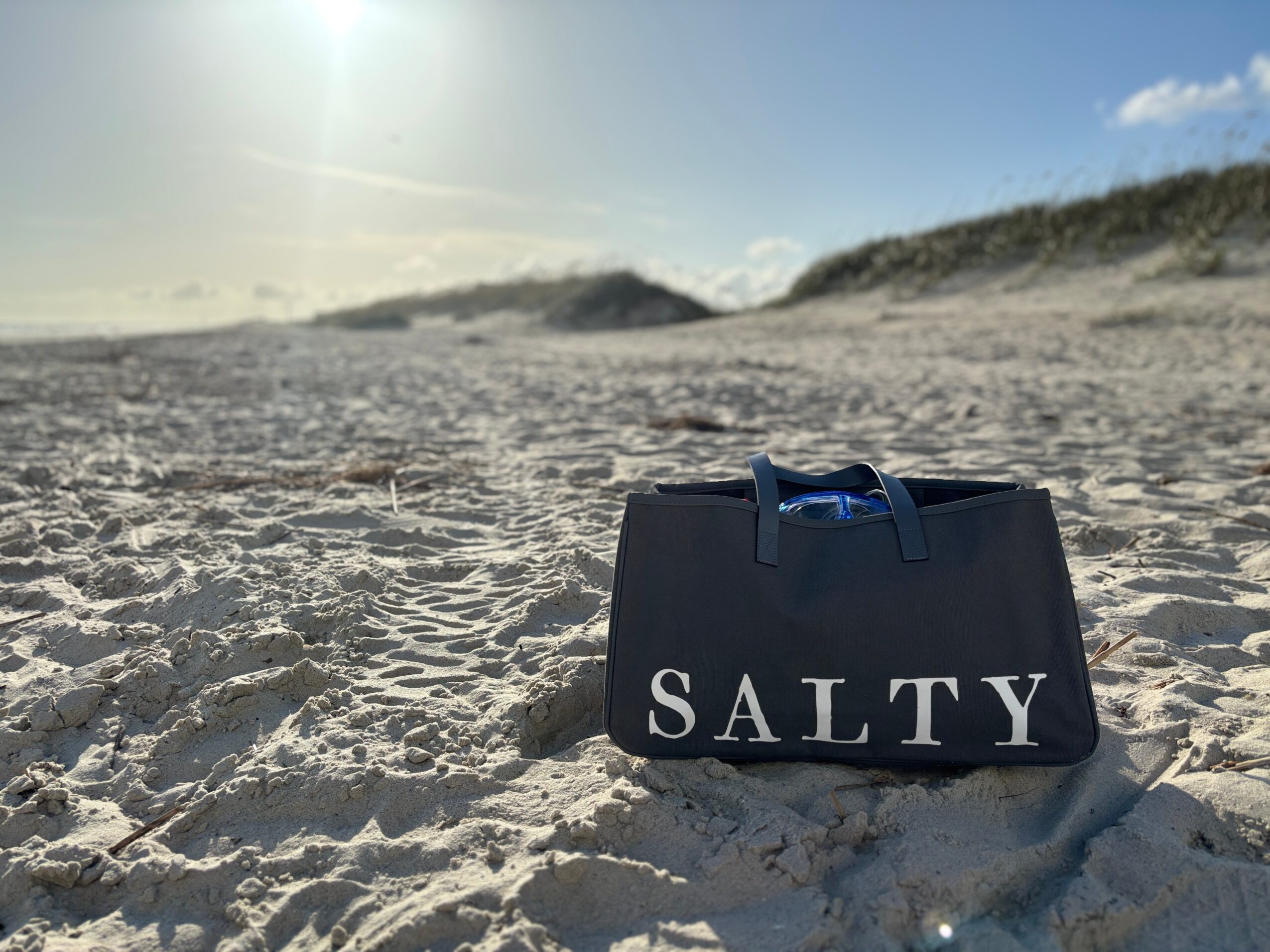 Black bag with white text reading SALTY on sandy beach, dunes in background, bright sky, sun shining.