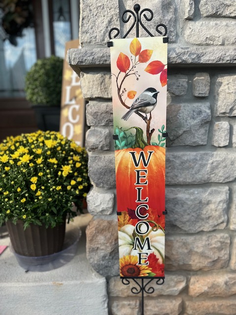 Vertical welcome sign with a bird and colorful leaves, mounted on a stone wall, with a potted yellow flower nearby.