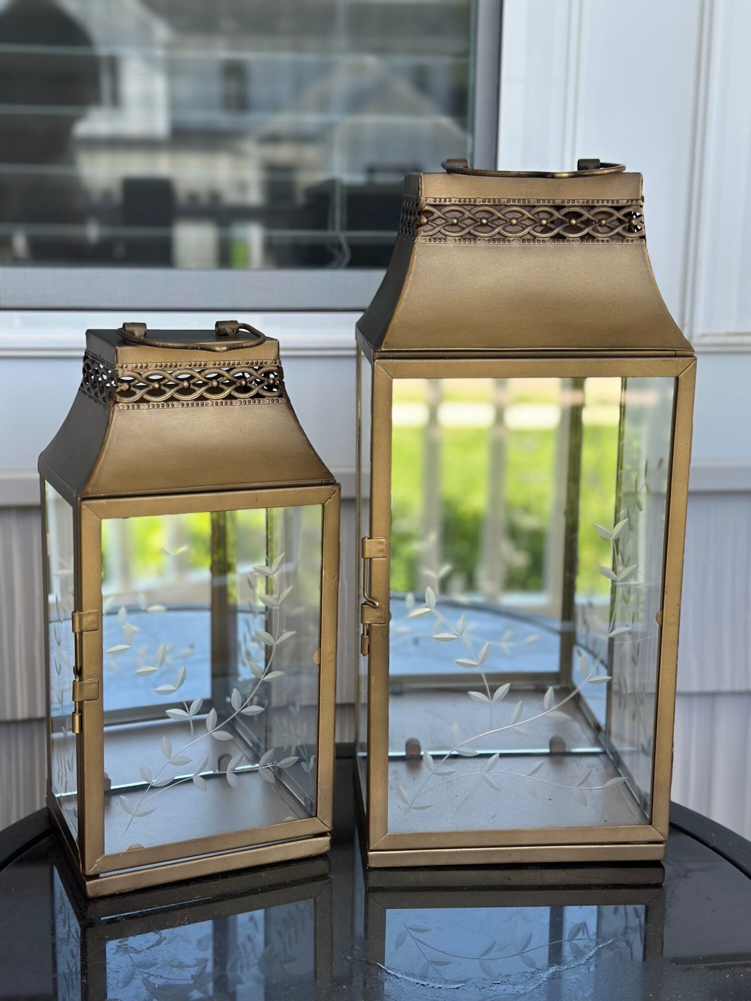 Two glass lanterns with metal frames and decorative tops, placed on a black surface near a window with a garden view.