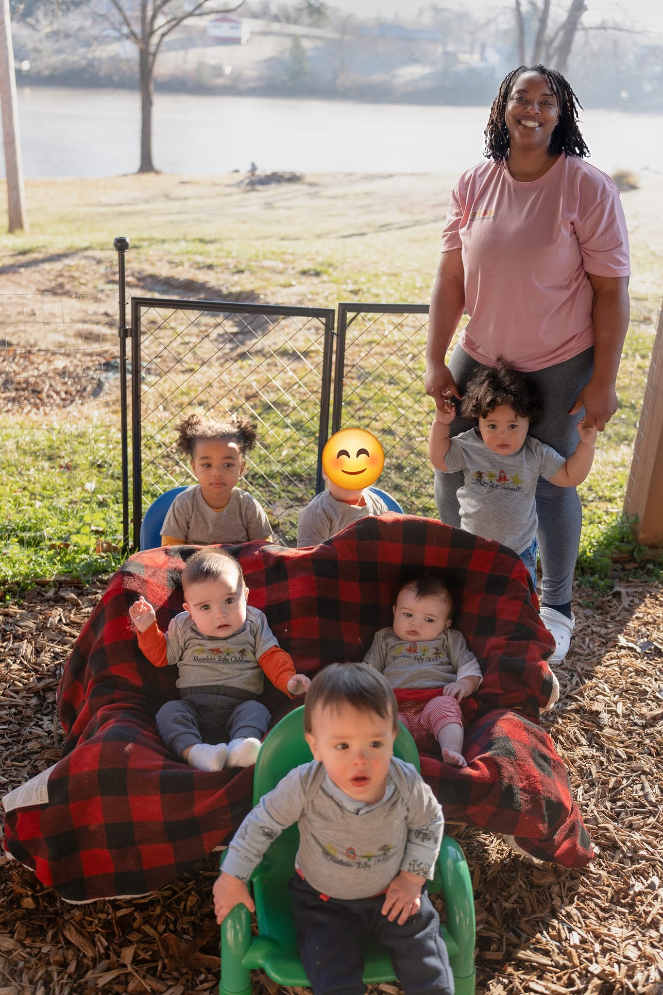 Group of five children and one adult outdoors near a lake, with a red and black blanket-covered bench, trees, and grass.