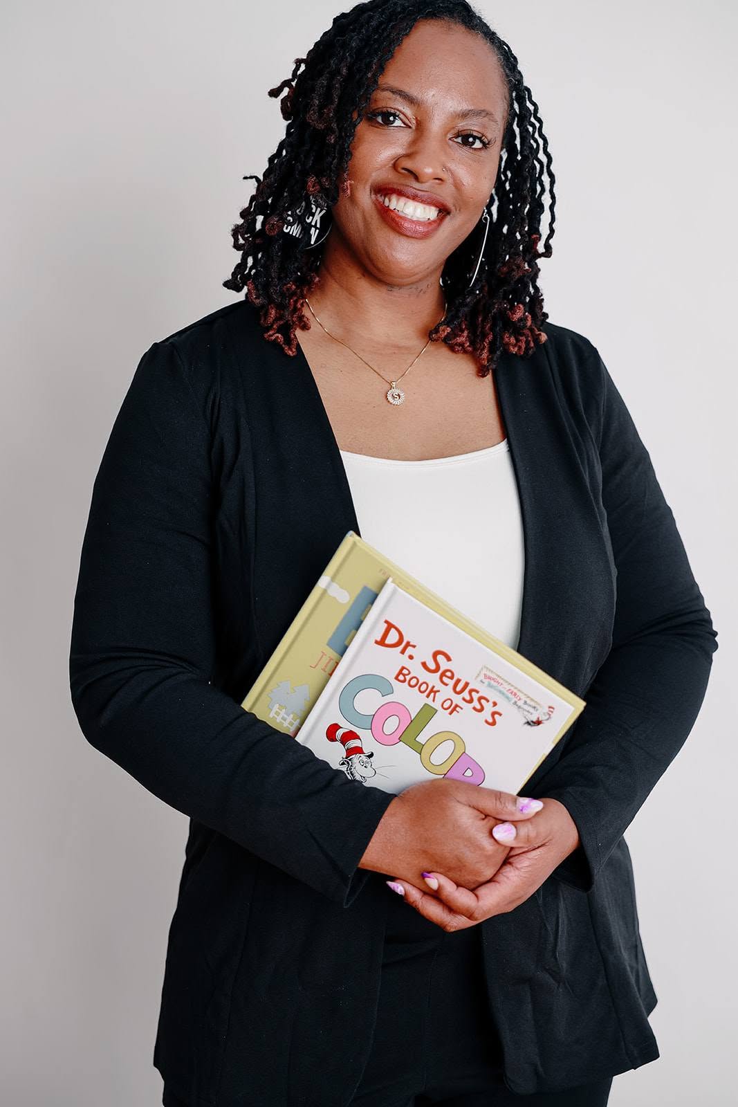 Woman with curly hair holding a book, smiling, wearing a black cardigan and white top, against a plain background.