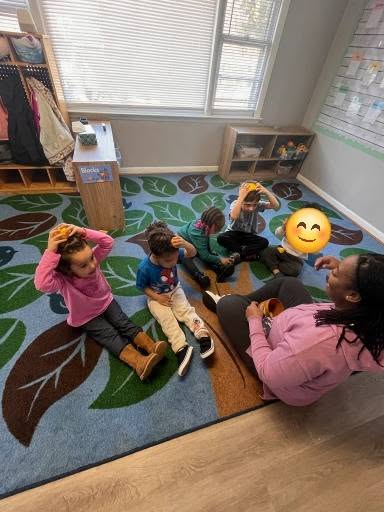 Children sitting on a colorful rug with a teacher, engaging in an activity, in a classroom with windows and shelves.