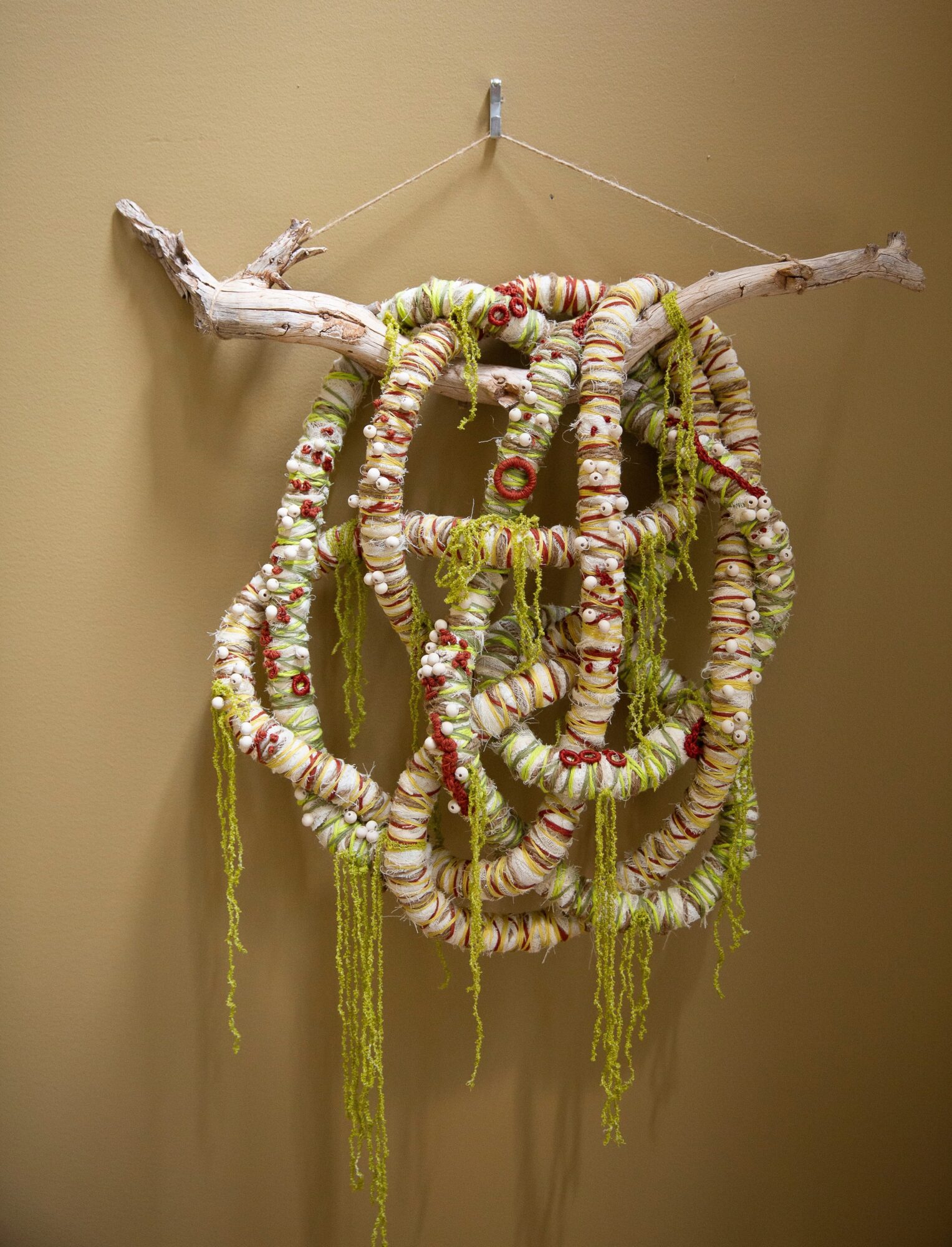 Decorative hanging wreath made of fabric, beads, and yarn on a beige wall, with a branch at the top.