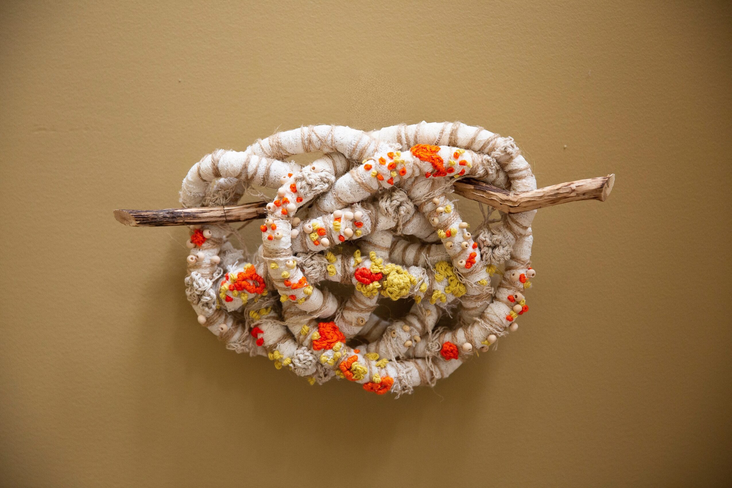 Coiled fabric with orange and yellow accents wrapped around a stick, mounted on a beige wall.