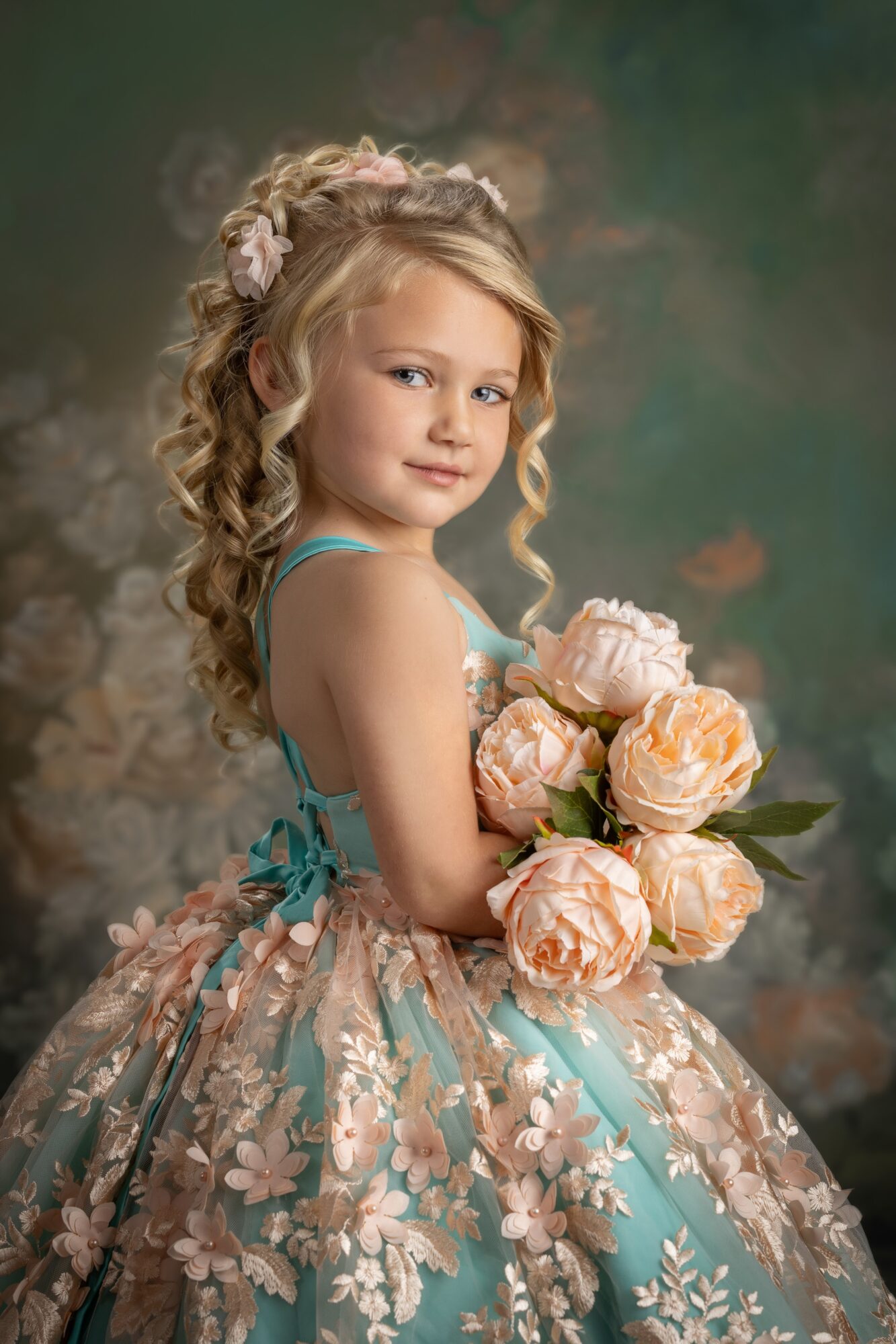 Young girl with curly blonde hair holding a bouquet of pink roses, wearing a floral dress, looking at the camera.