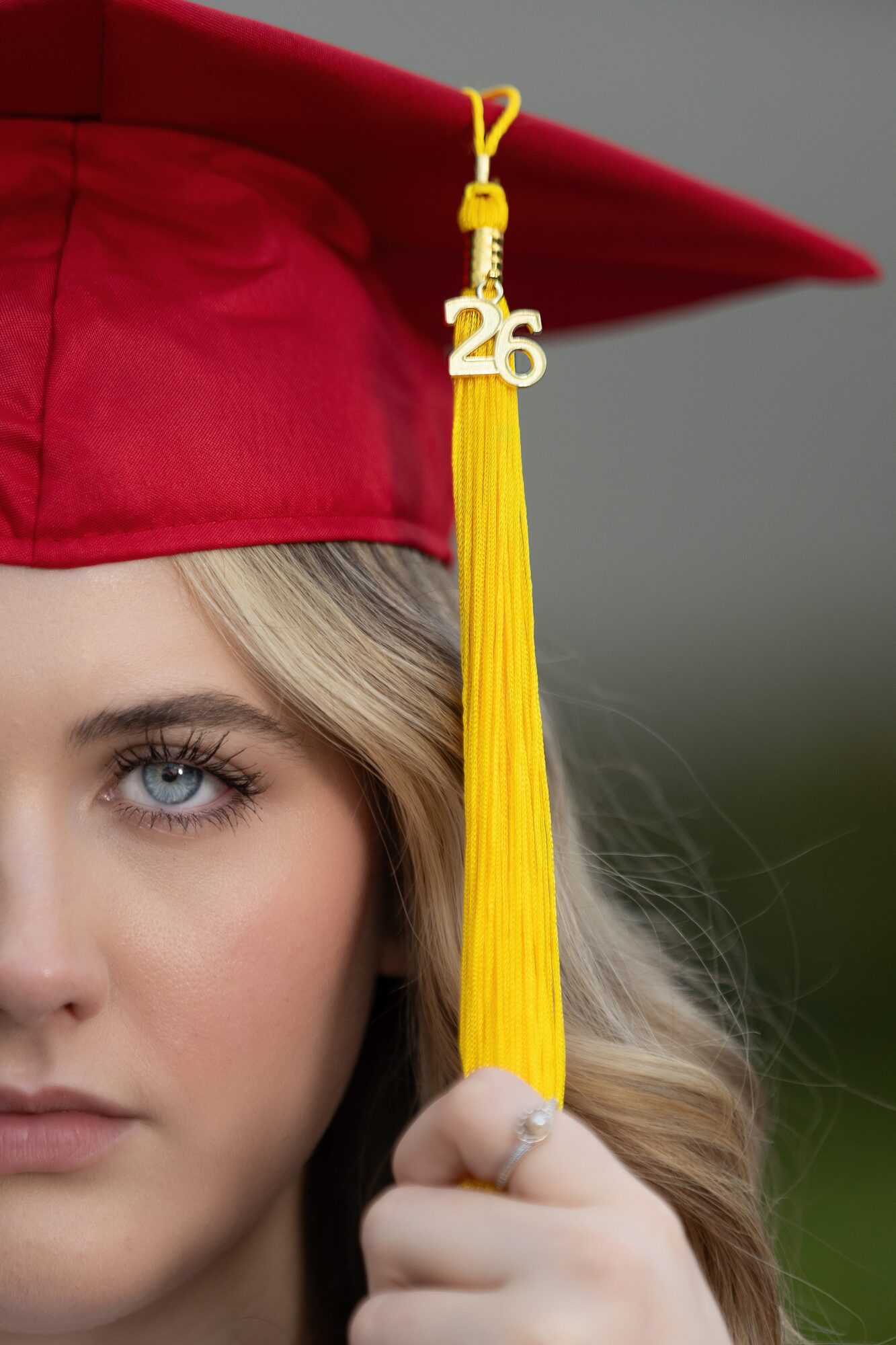Close-up of a woman wearing a red graduation cap with a yellow tassel and a '26' charm, holding the tassel.