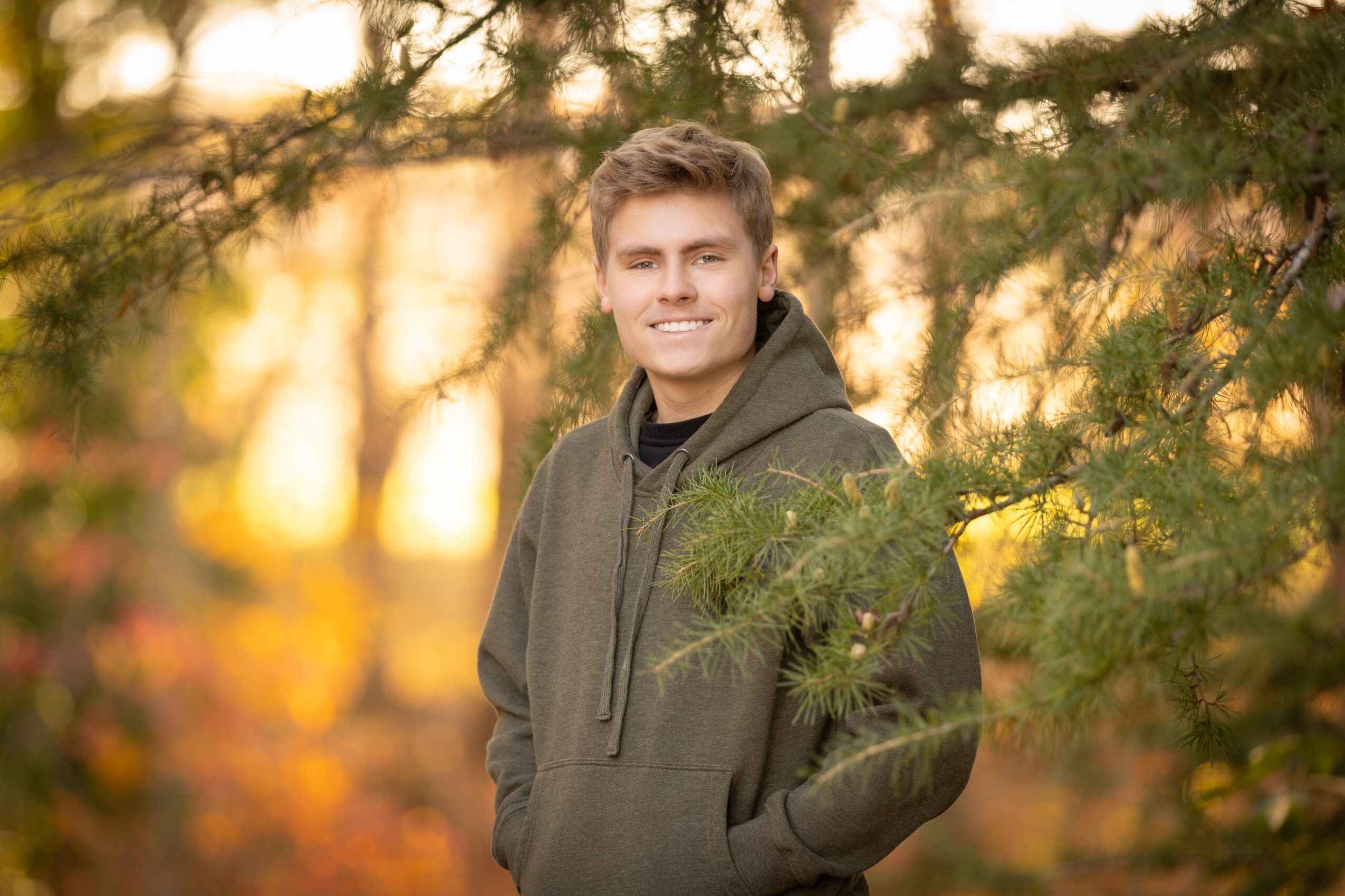 Young man smiling outdoors in autumn, surrounded by trees with fall foliage, wearing a dark hoodie.