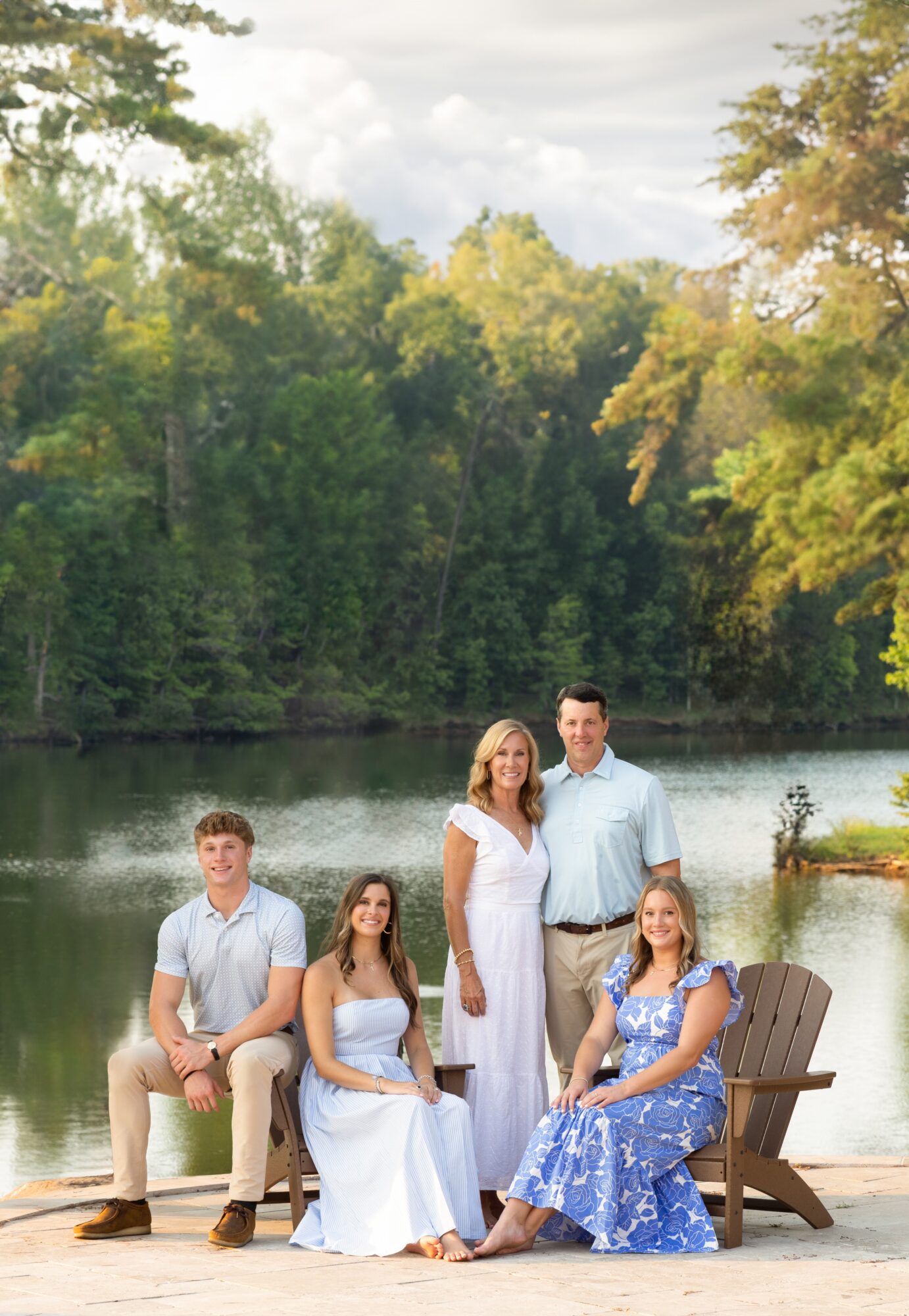 Four people sitting and standing near a lake with trees in the background, two women and two men, outdoors.
