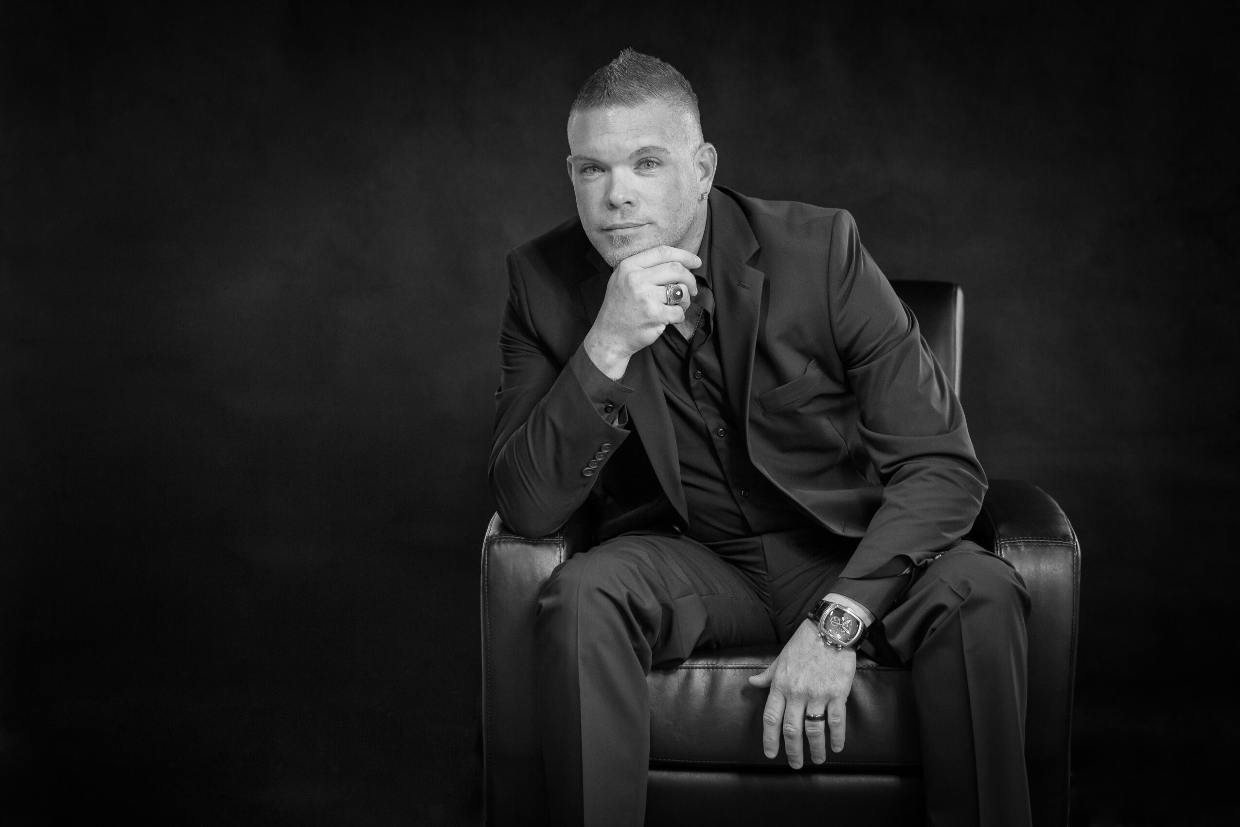 Man in suit sitting on a chair with hand on chin, black background, black and white photo.