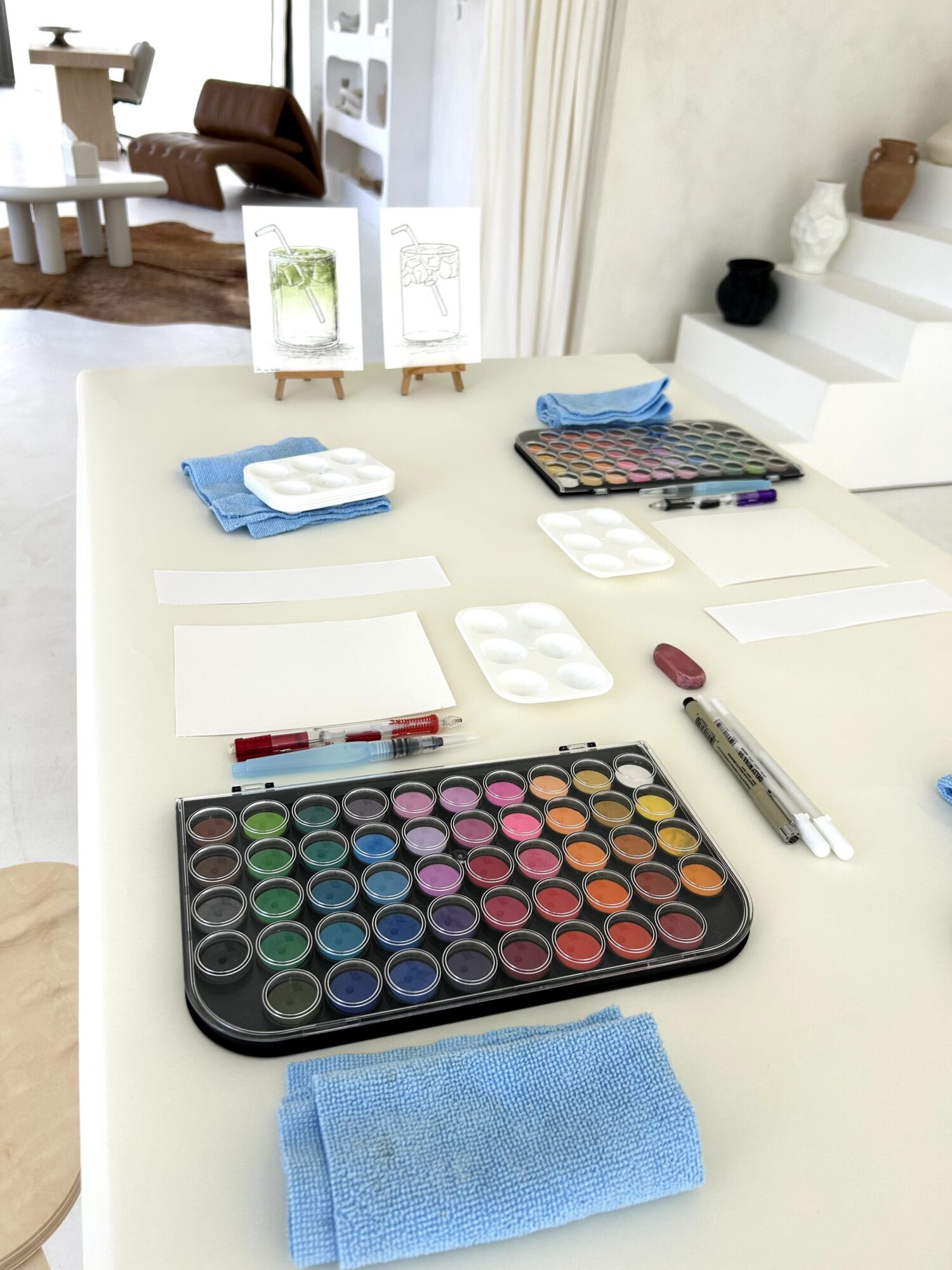 Art supplies including watercolor paints, brushes, paper, and towels on a table in a bright room.