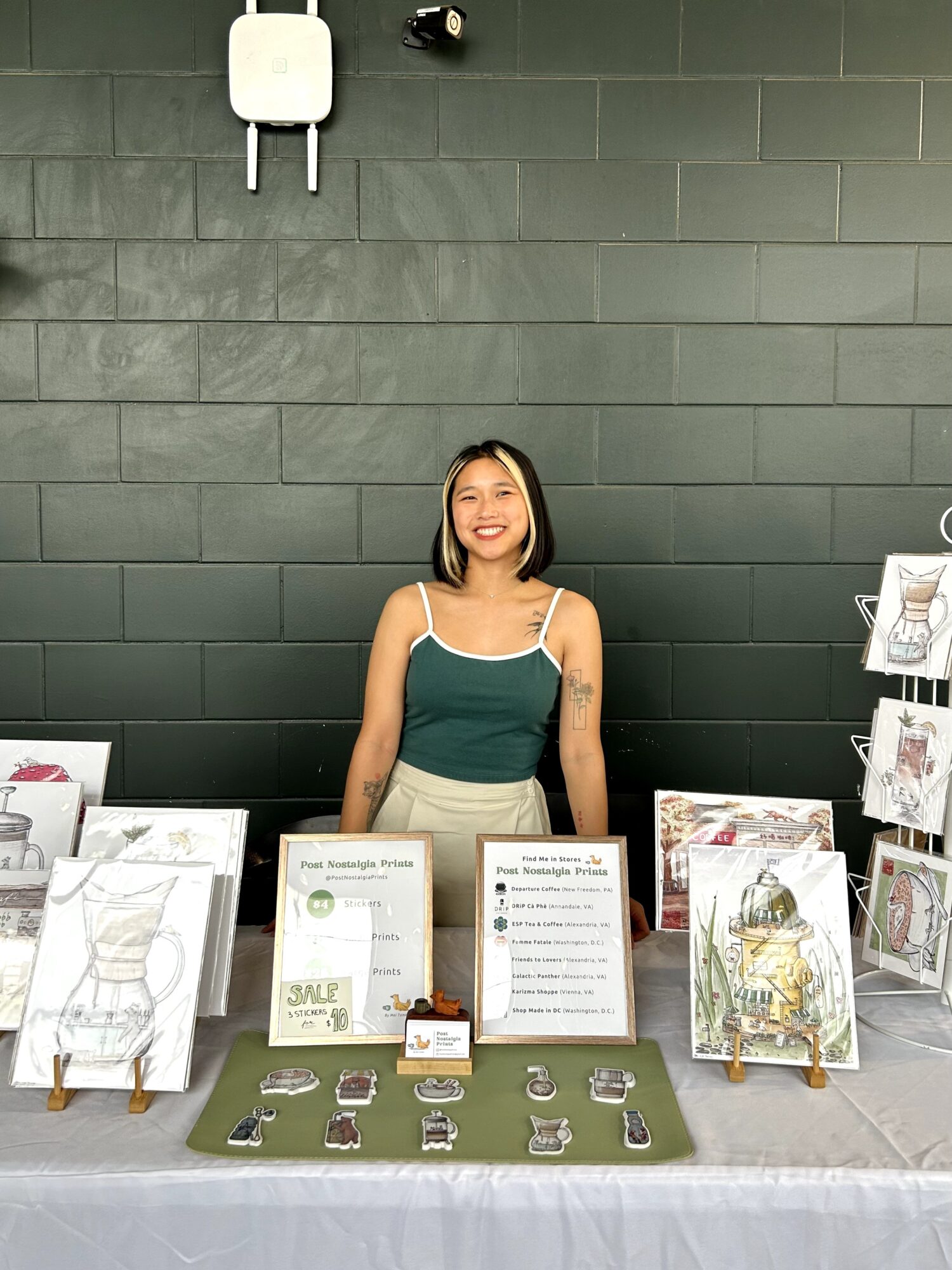 Young woman standing behind a table with jewelry displays and signs, smiling, against a dark green wall.