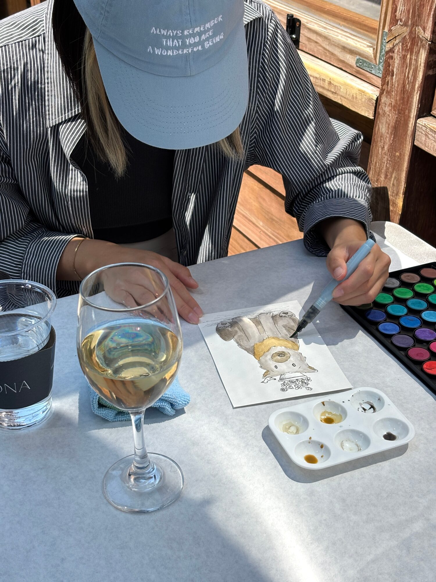 Person painting on paper with watercolor set, glass of white wine, and water glass on table outdoors.