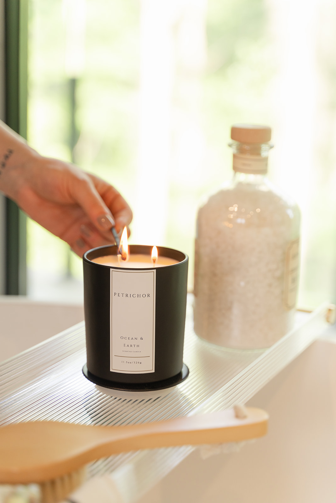 Person's hand lighting a black candle on a white surface with a glass bottle nearby, sunlight through window in background.