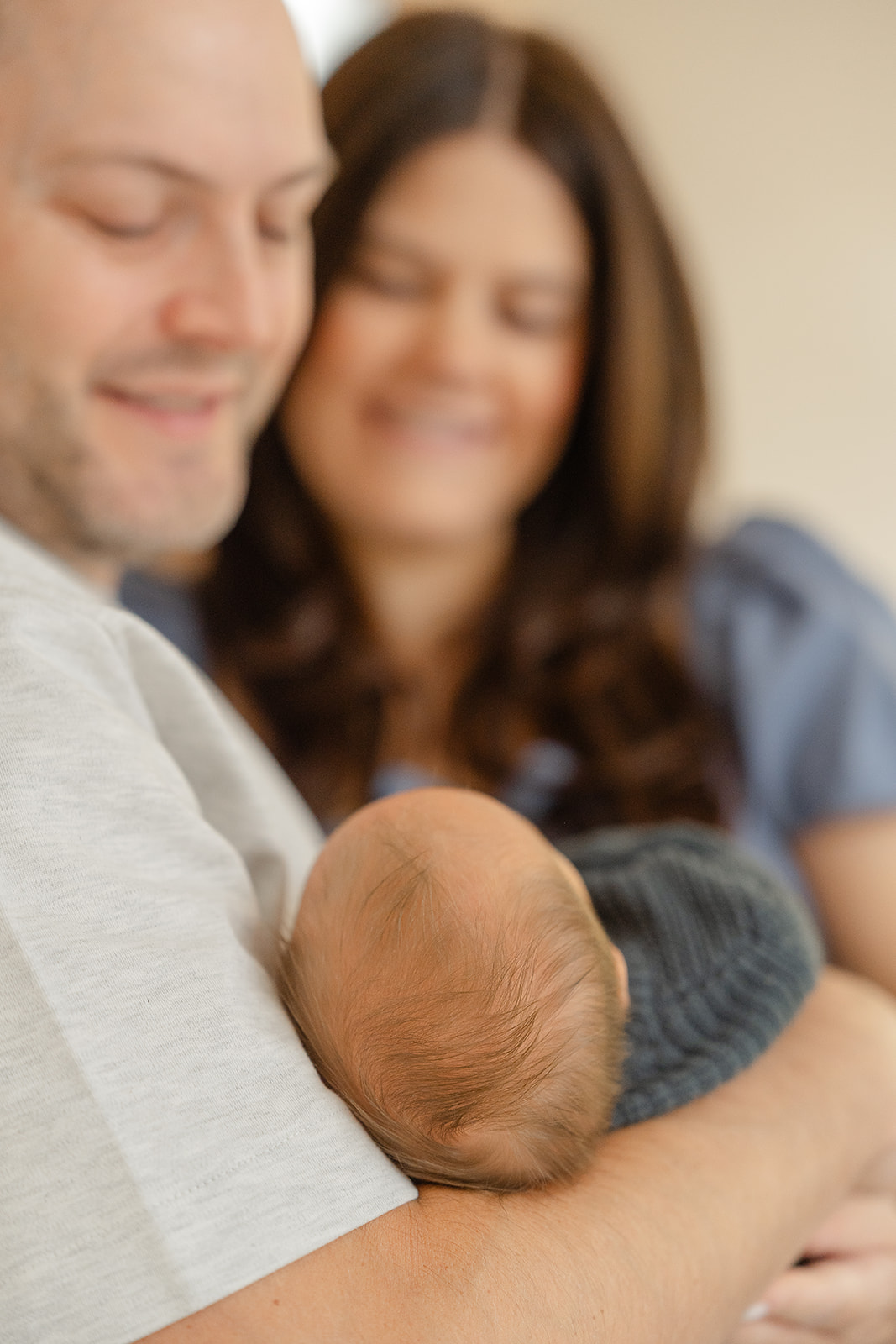A man and woman hold a newborn baby, smiling and looking at the baby, with a neutral background.