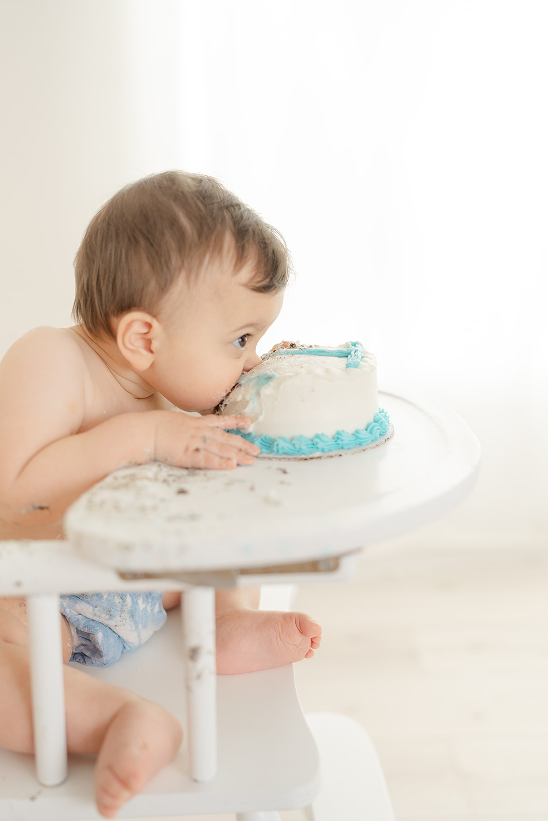 Baby leaning over a table to eat birthday cake with blue icing, sitting in a high chair, light background.