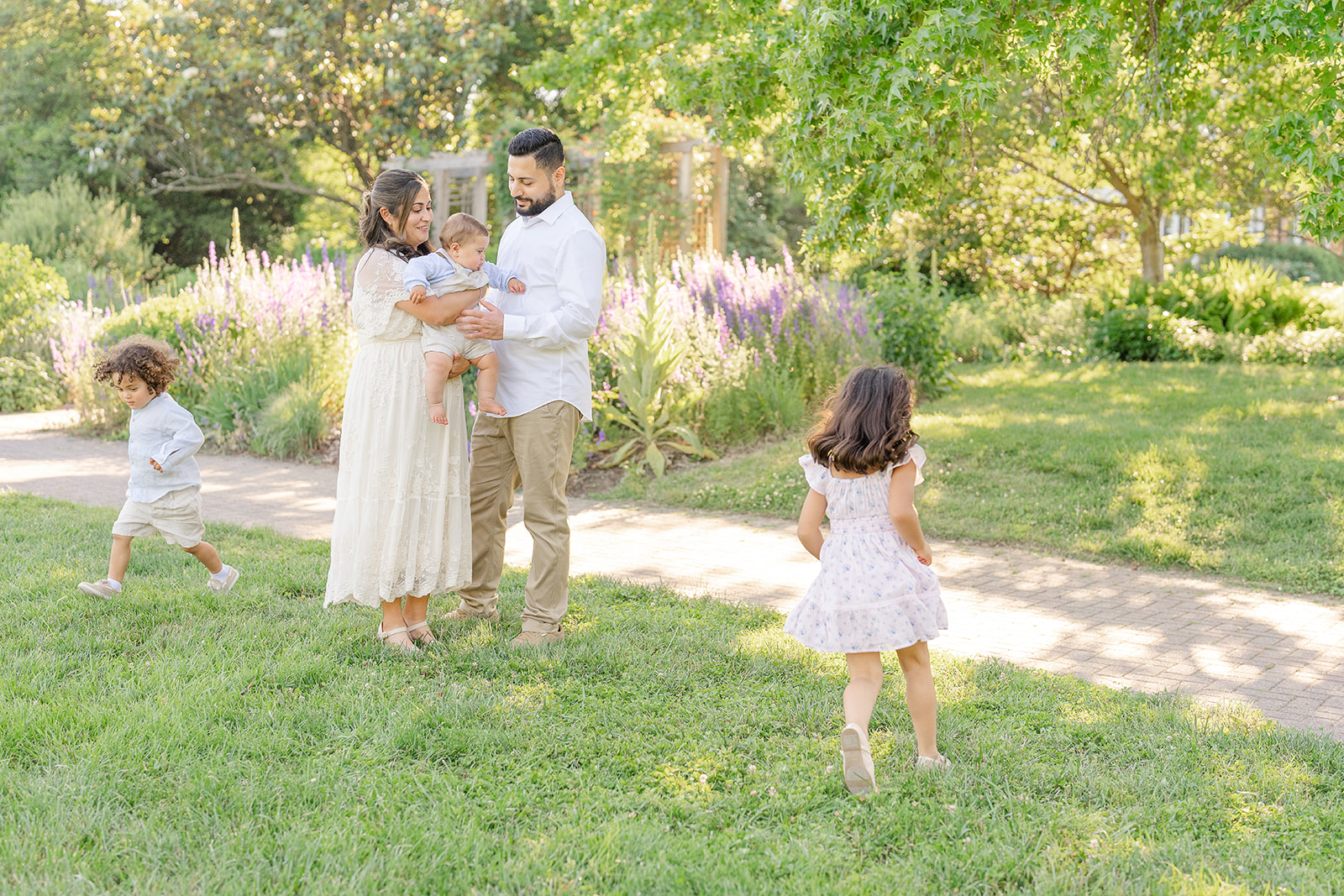 Family with two children in a park, trees and flowers in background, children playing on grass and path.