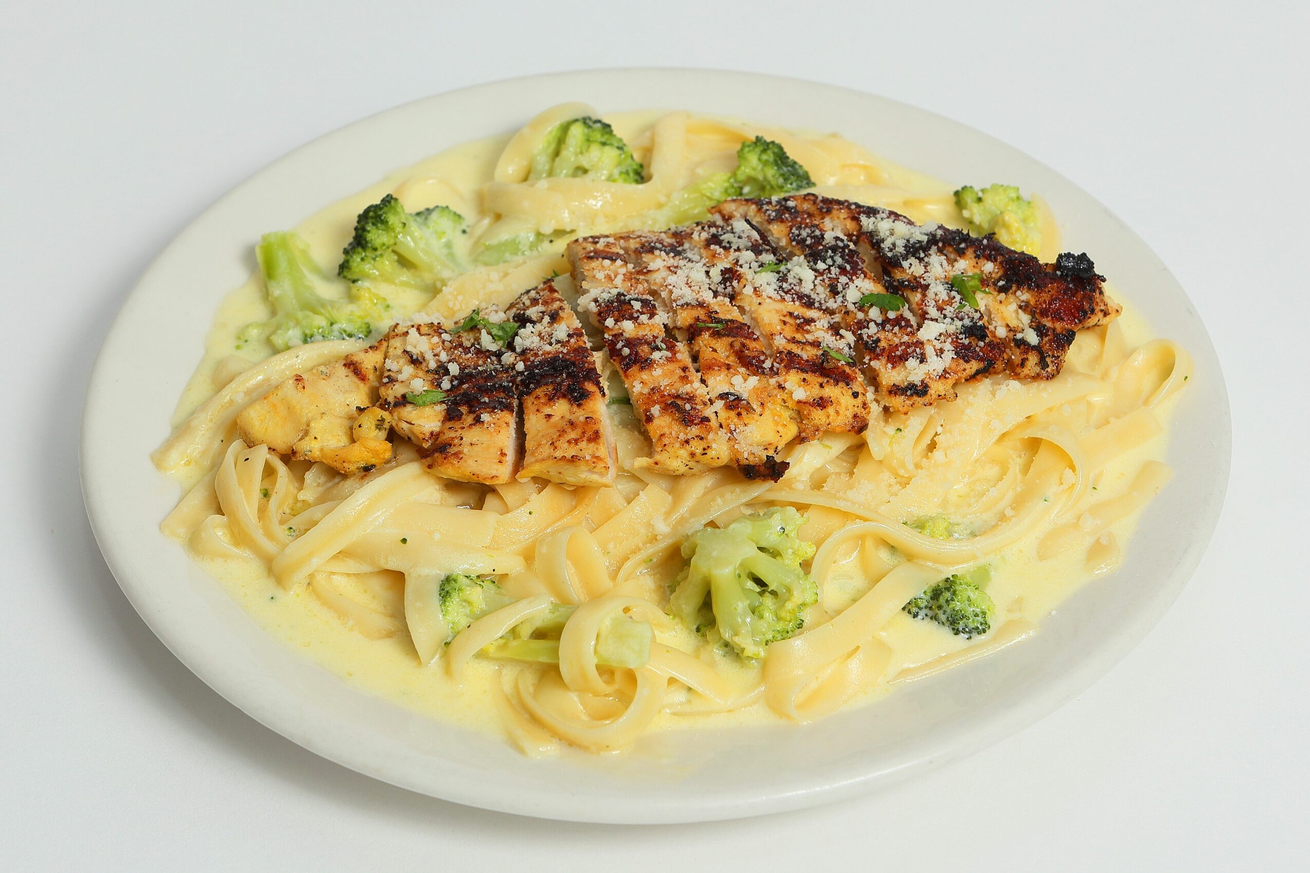 Plate of pasta with grilled chicken and broccoli, topped with grated cheese, on a white background.