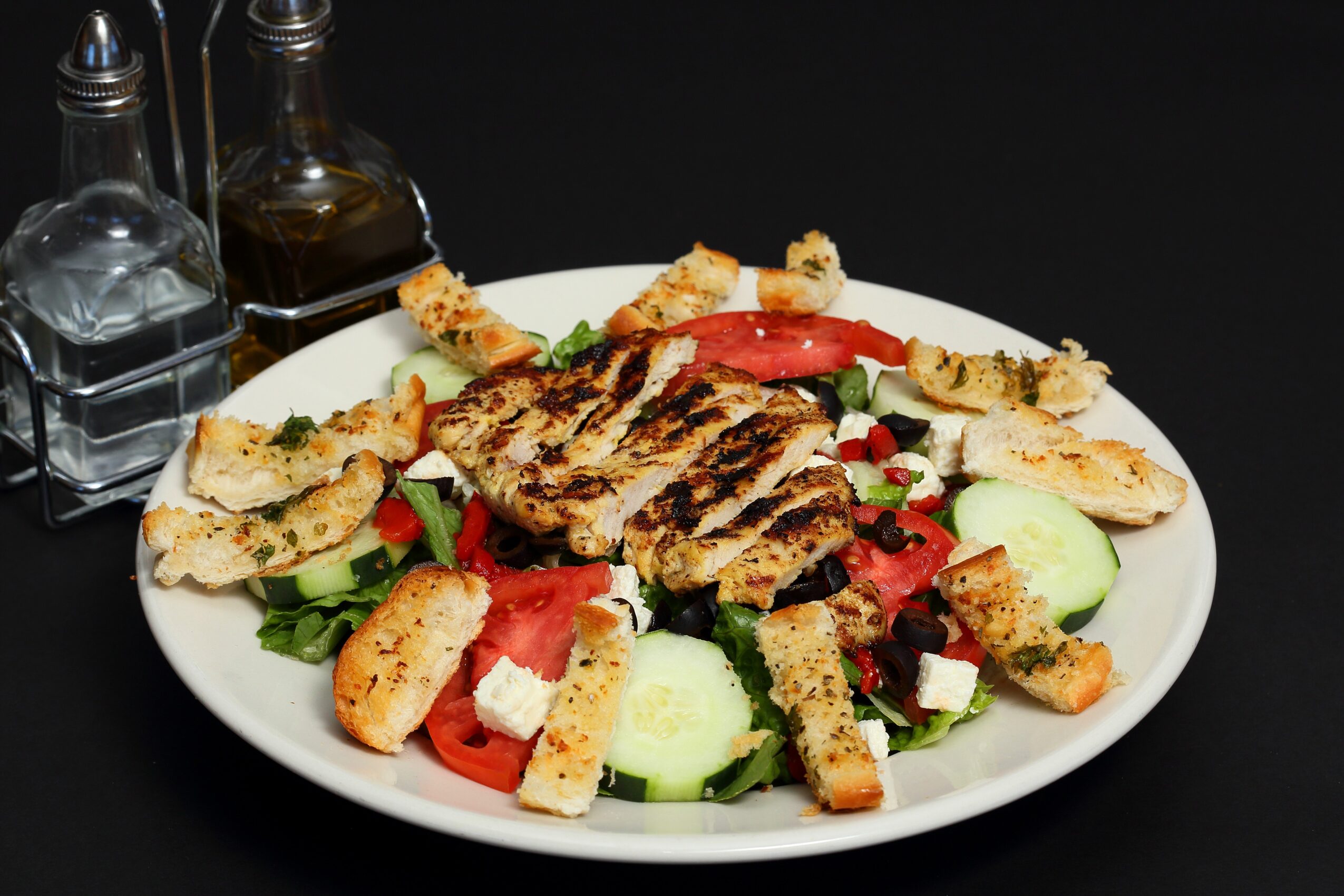 Grilled meat with cucumber, tomato, feta cheese, and breadsticks on a white plate, with salt and oil bottles in the background.