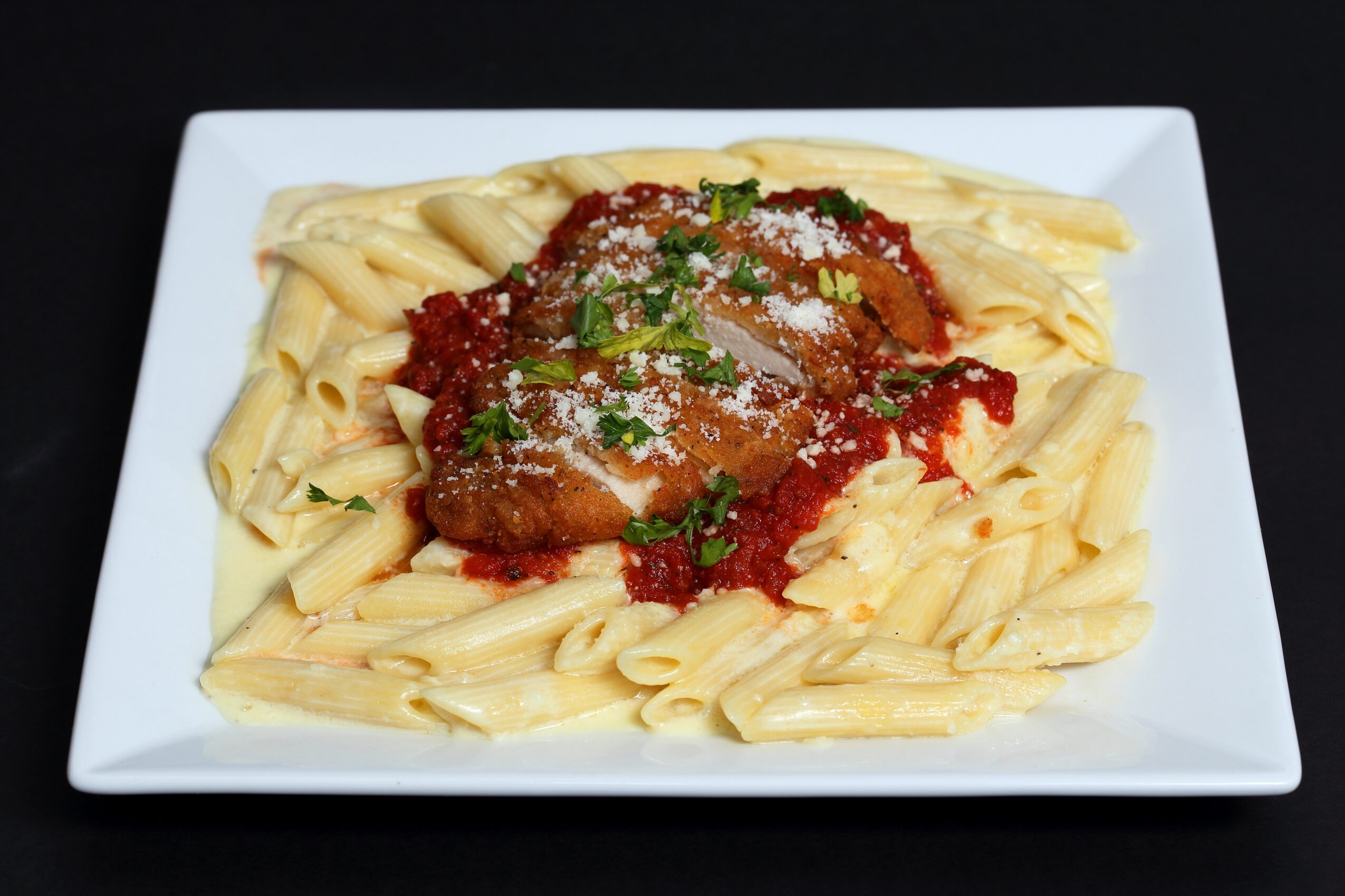 Plate of pasta with meat sauce, garnished with parsley and grated cheese, on a white square dish against a black background.