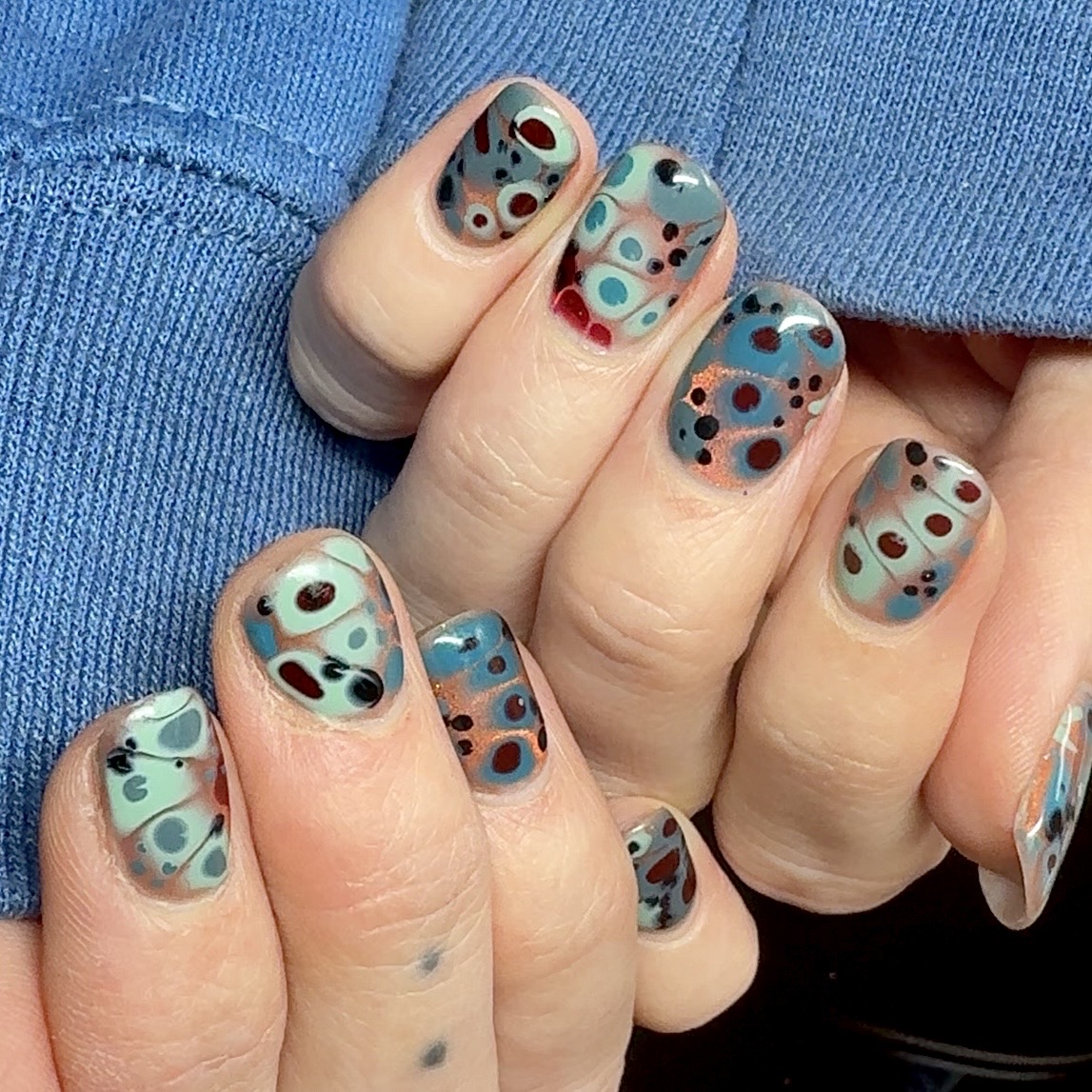 Hand with colorful polka dot nail art, blue sweater in background, nails are short and decorated with various colored dots.