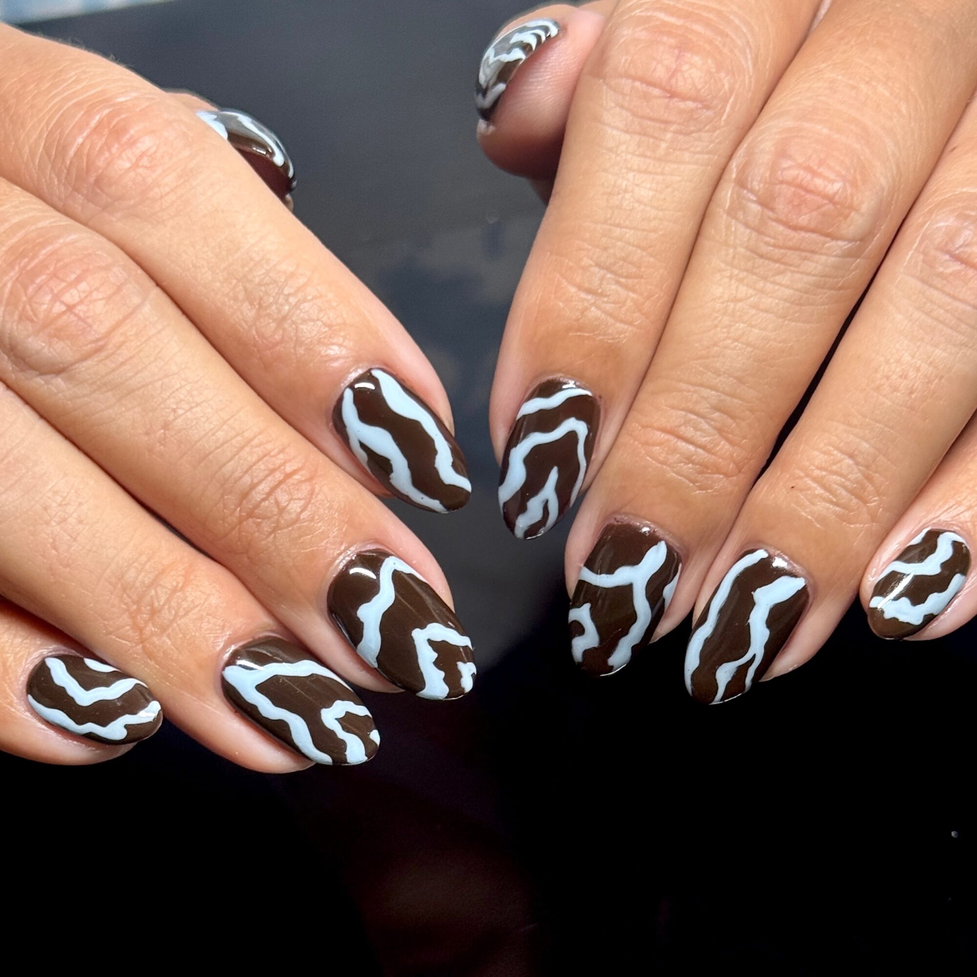 Hands with nails painted in a black and white wavy pattern, against a dark background.