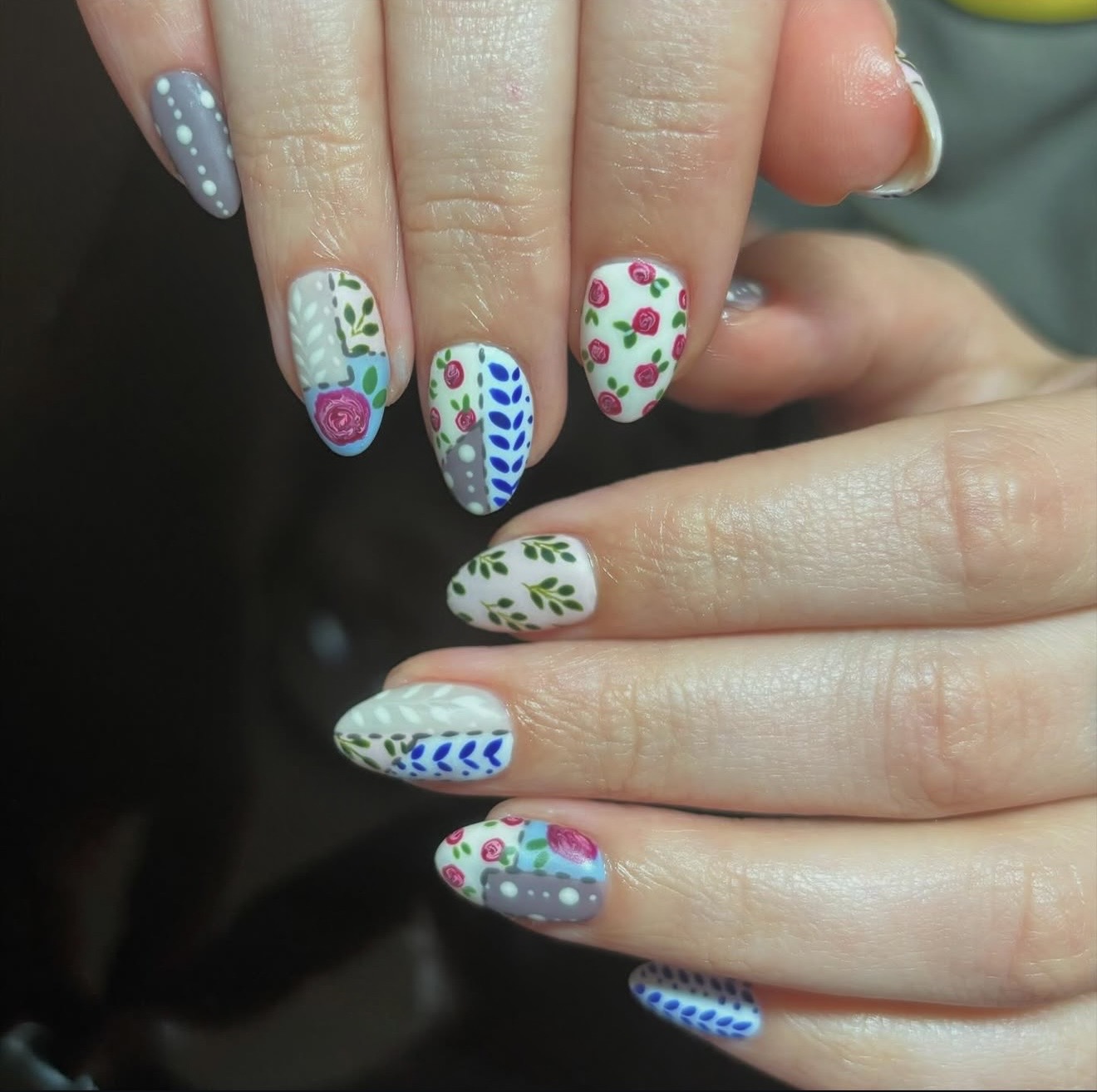 Close-up of hands with colorful floral and dotted nail art on fingernails, some with white backgrounds and others with gray backgrounds.