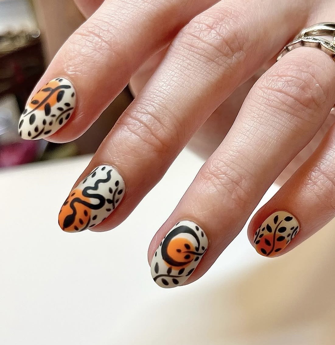 Four fingers with colorful, abstract nail art designs, including orange, black, and white patterns, on a person's hand.