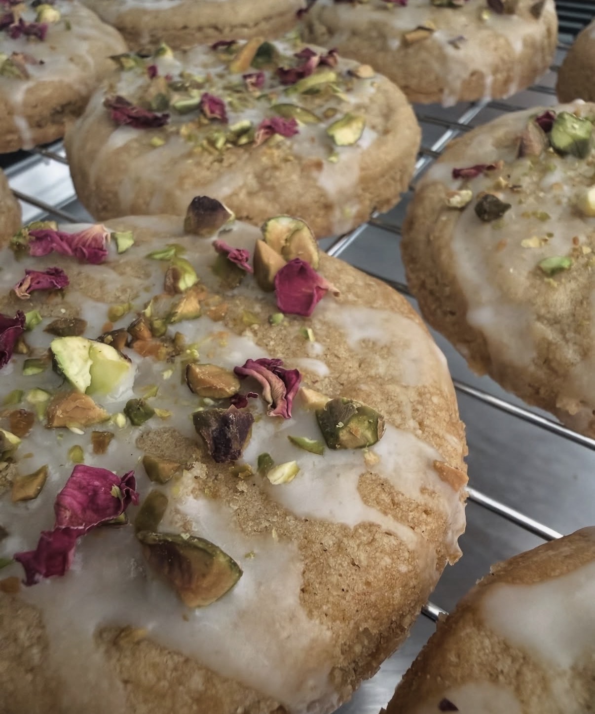 Close-up of heart-shaped cookies topped with white icing, pistachios, and pink rose petals, on a wire rack.