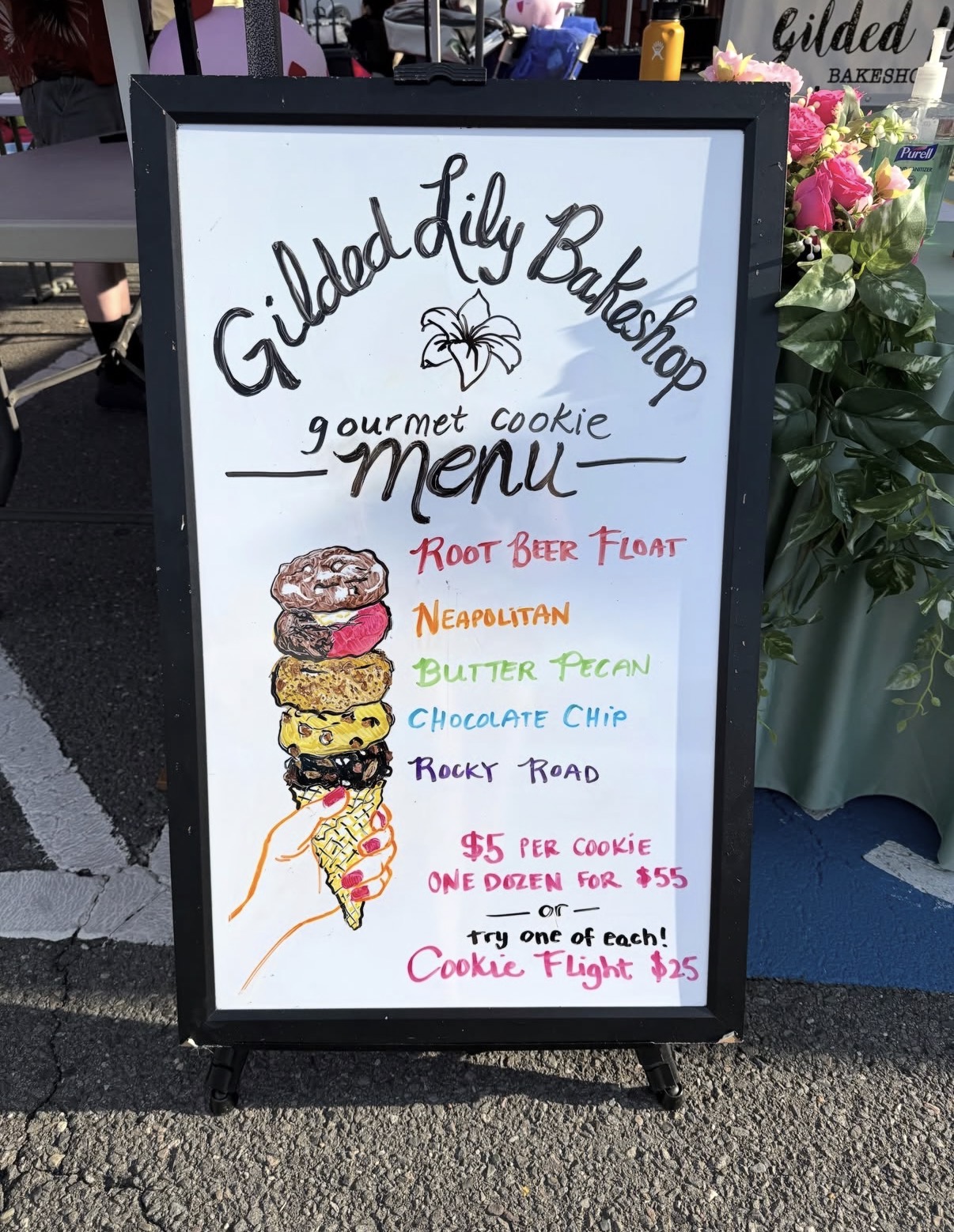 Whiteboard menu with colorful handwritten text and a drawing of a cookie with ice cream and toppings, outdoors on pavement.