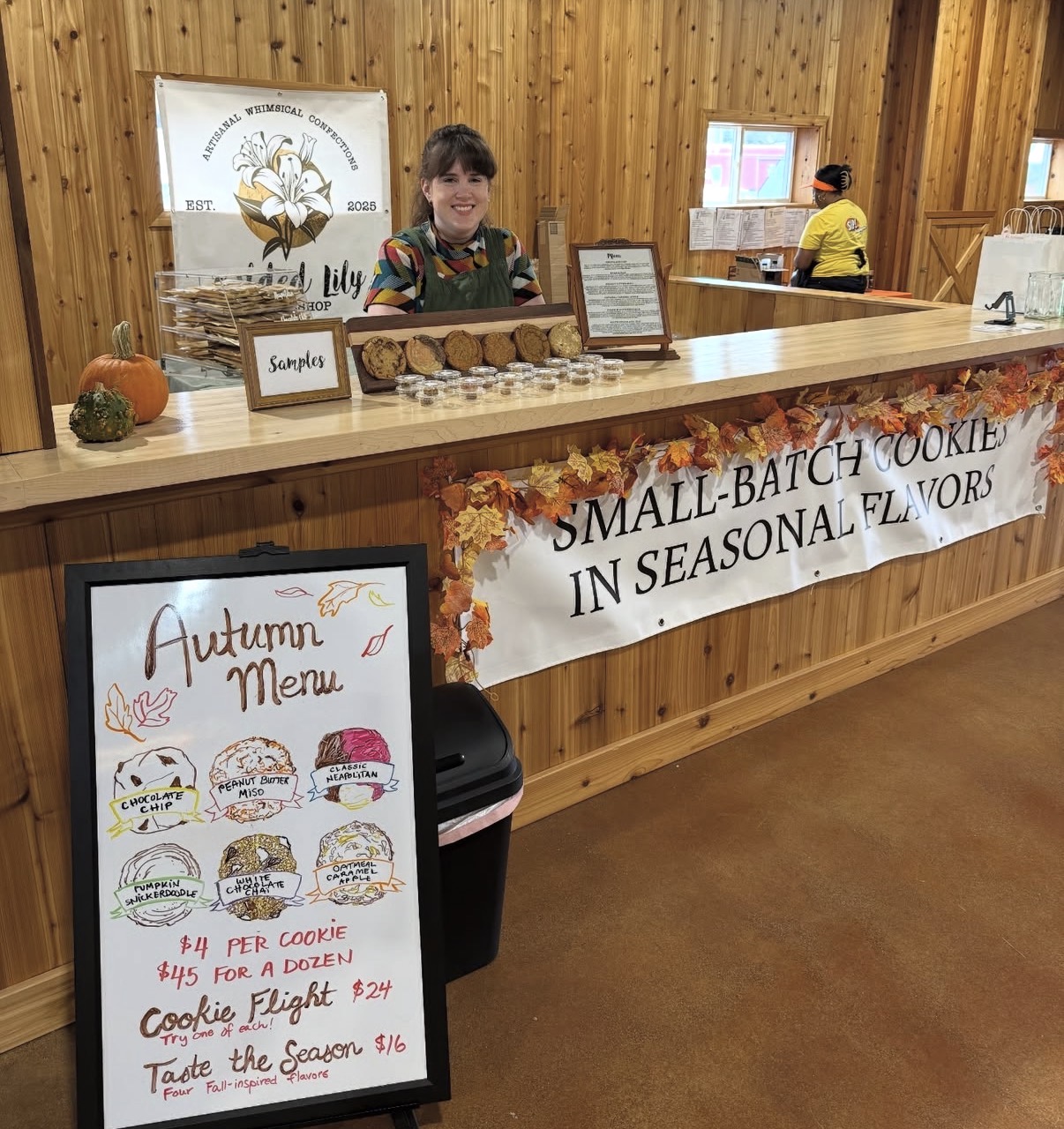 Person behind a wooden counter with seasonal cookie display, autumn menu sign, and a banner advertising small-batch cookies in seasonal flavors.