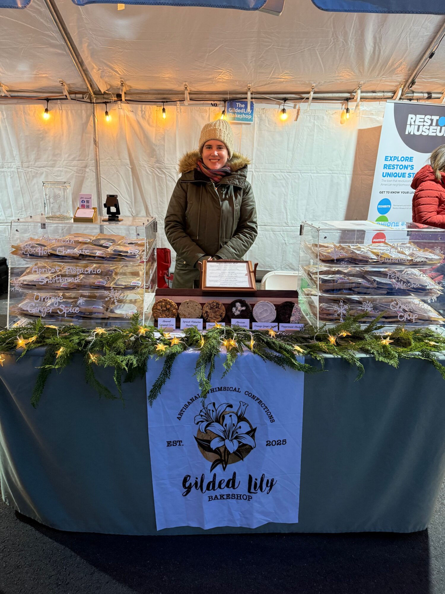 Person standing behind a table with food and a sign, inside a tent with lights, at an event.
