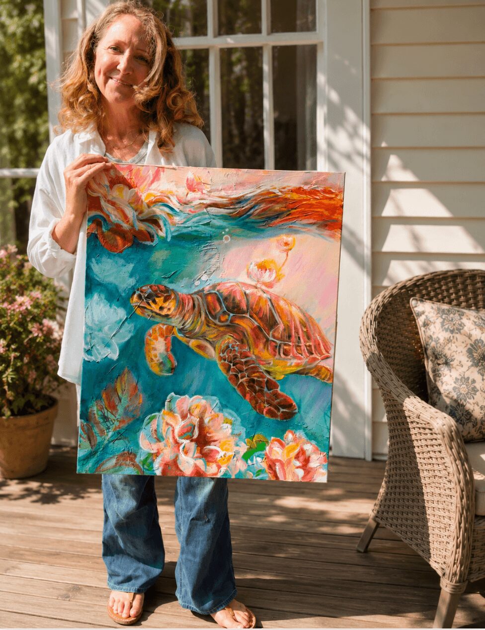 Woman holding a colorful painting of a turtle swimming among flowers outdoors on a porch.
