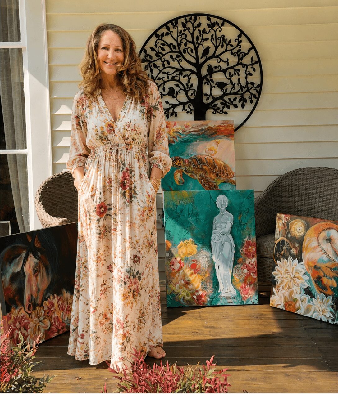 Woman in a long floral dress standing indoors with colorful paintings and a decorative tree wall art behind her.