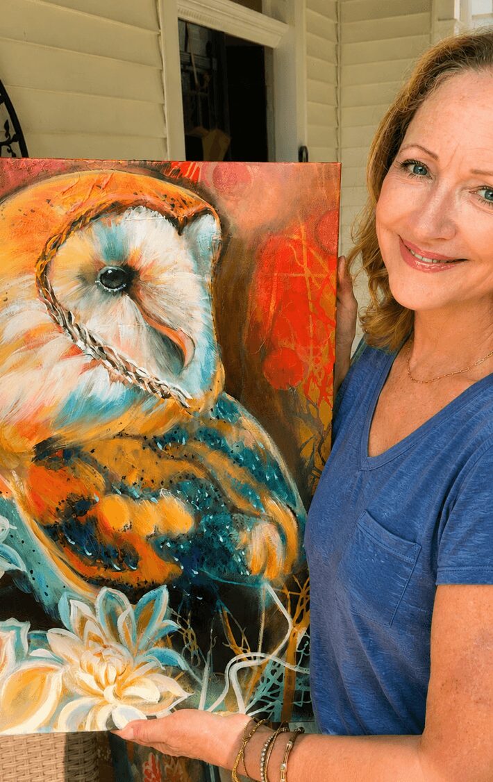 Woman holding colorful owl painting indoors, smiling, wearing blue shirt, with window and siding in background.