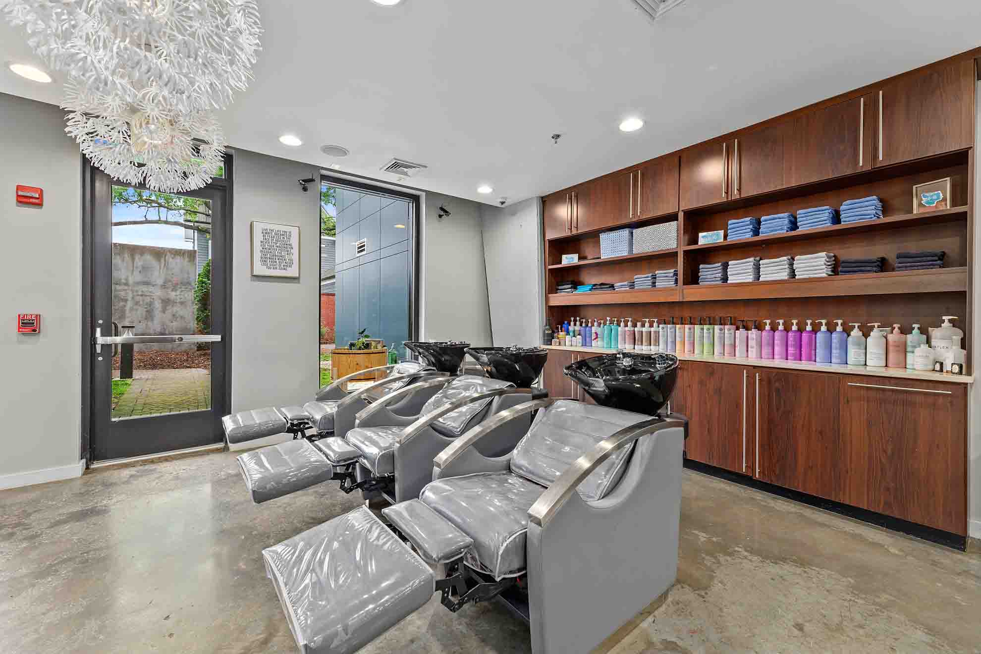 Salon with three shampoo chairs, wooden shelves with hair products, and a chandelier, near a glass door and window.
