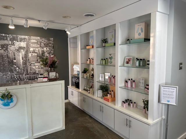 Interior of a store with shelves displaying skincare products, plants, and framed pictures, and a black-and-white cityscape mural on the wall.