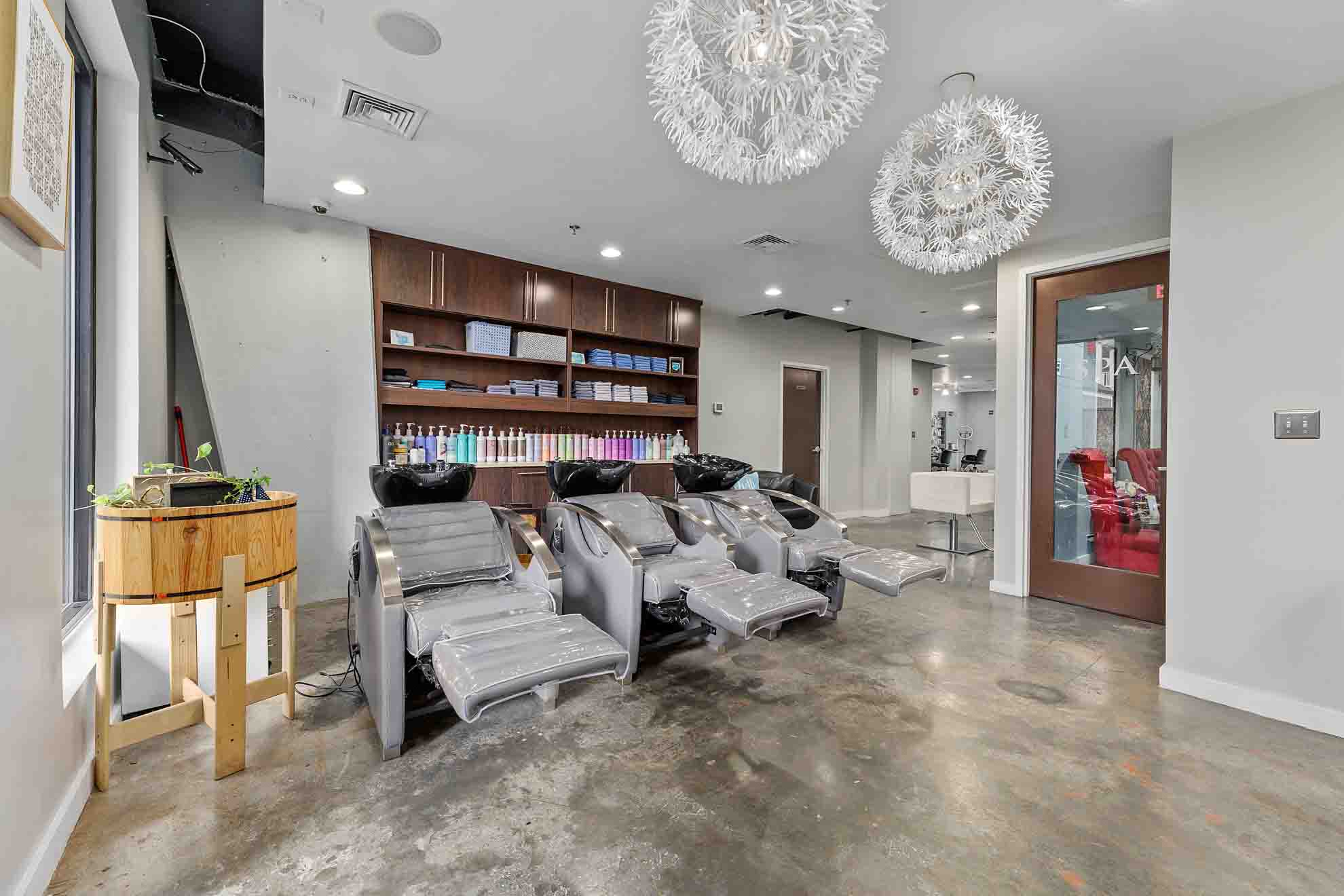 Salon with massage chairs, wooden shelves, decorative ceiling lights, and a glass door, with a polished concrete floor.