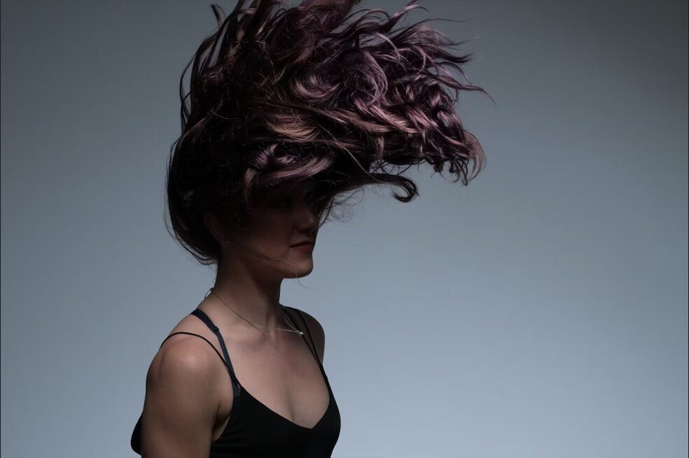 Person with curly hair flipping in the air, wearing a black top, against a plain background.