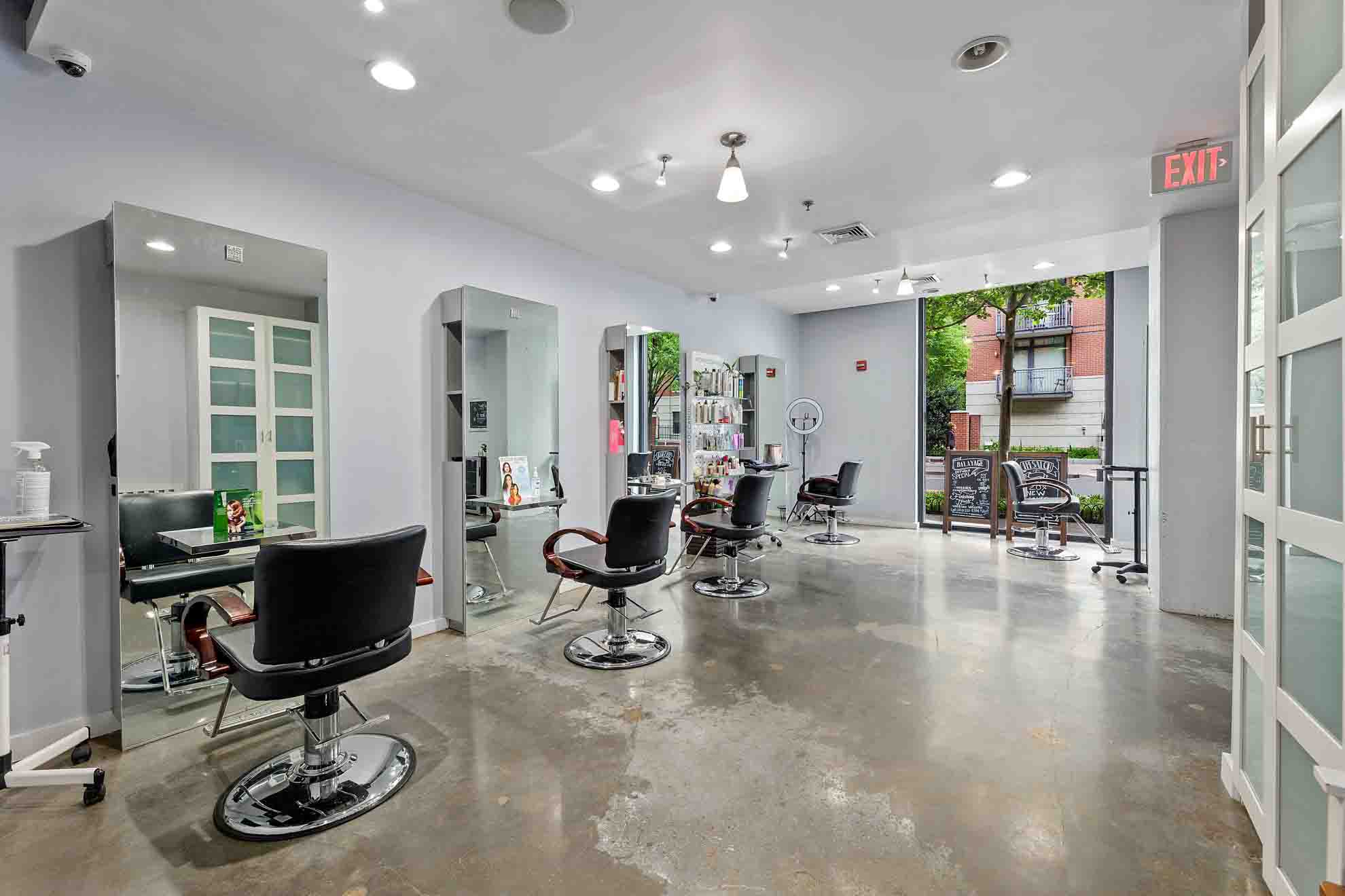Empty hair salon with chairs, mirrors, and styling stations, large windows, and a door leading outside, bright and clean space.