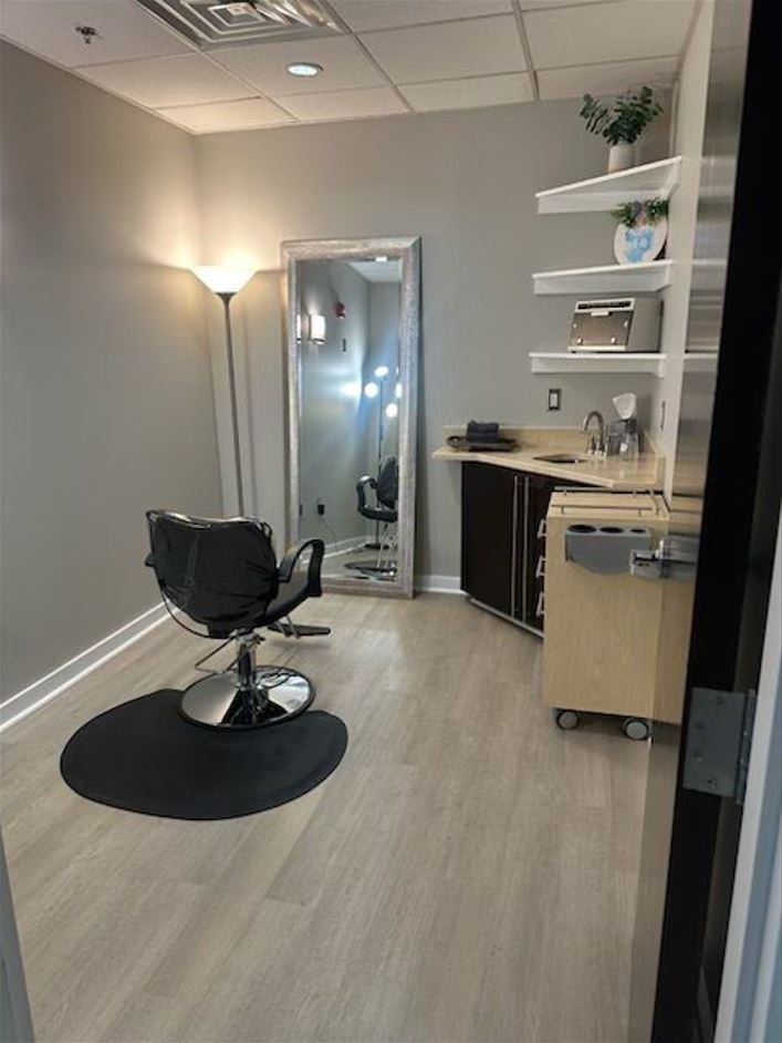 Salon with a styling chair, mirror, shelves, and a small cart, illuminated by a floor lamp in a neutral-colored room.