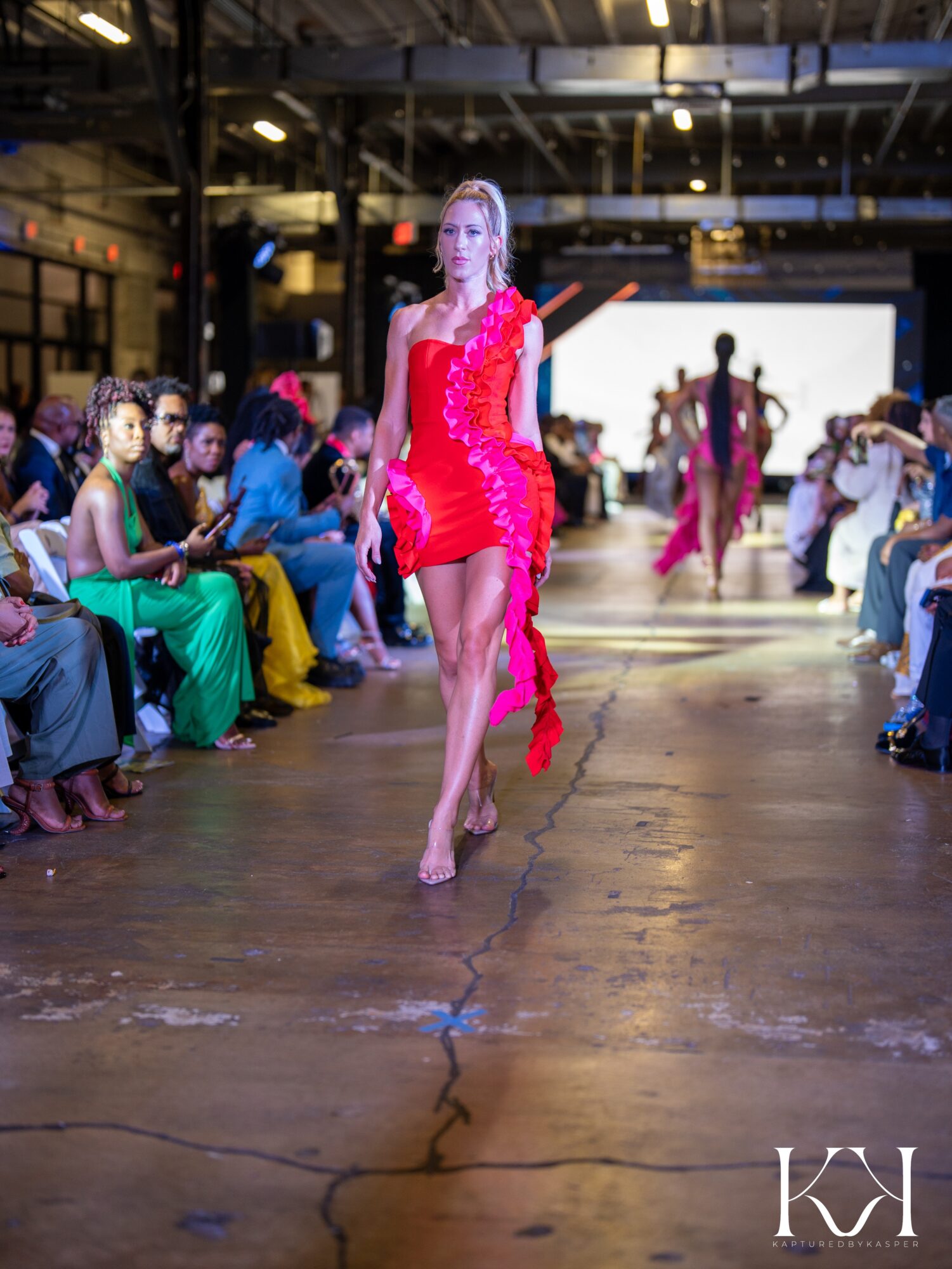 Model walking on runway in red dress with audience on both sides, industrial setting, bright lighting, large screen in background.