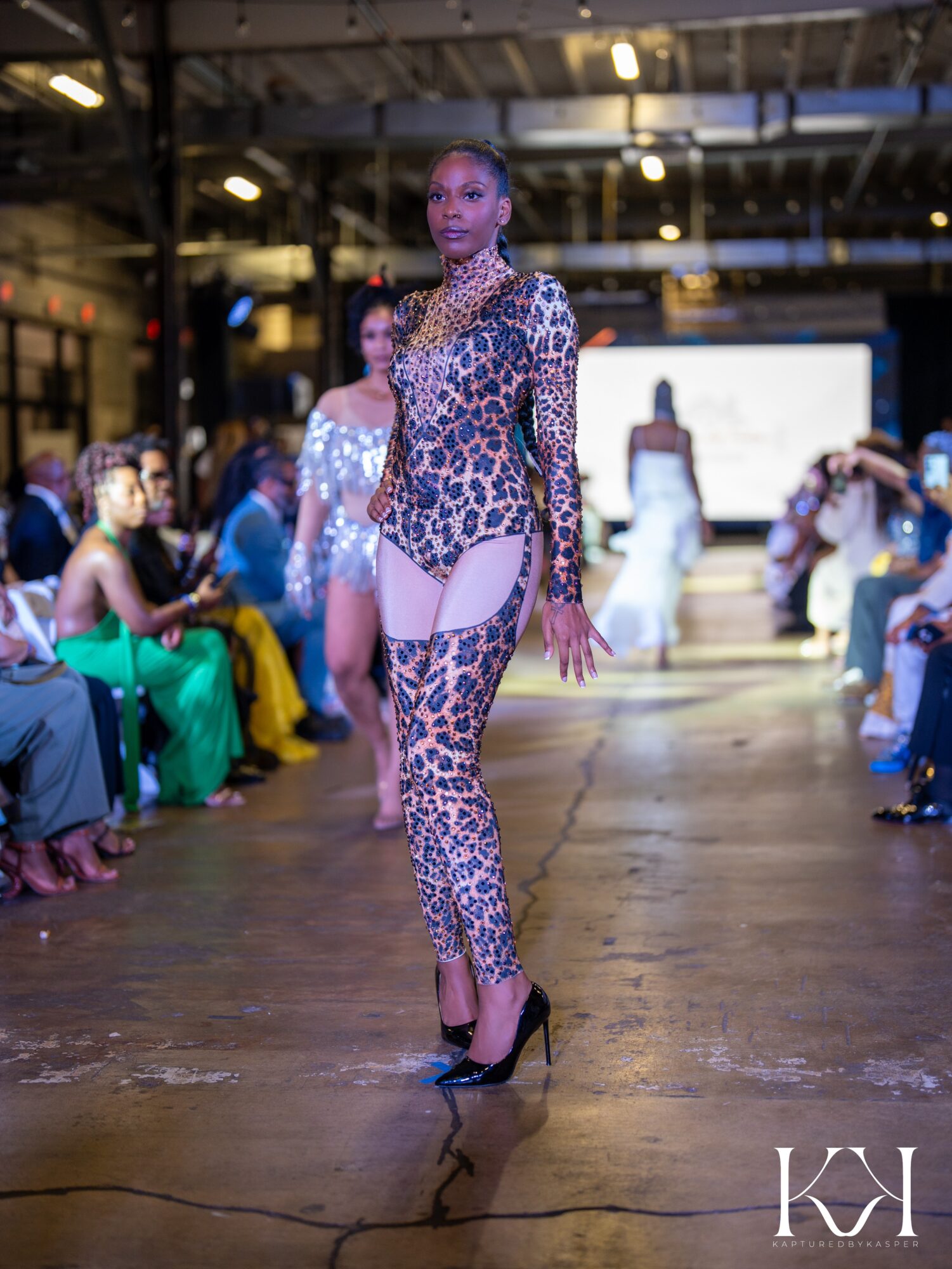 Model walking on runway in leopard print outfit, audience seated on both sides, industrial setting, bright lighting.