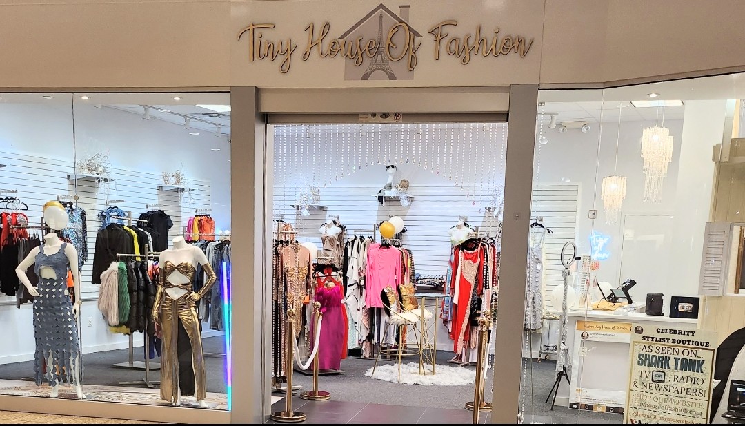 Clothing store entrance with mannequins displaying dresses and outfits, store name above, interior visible through glass, no people outside.