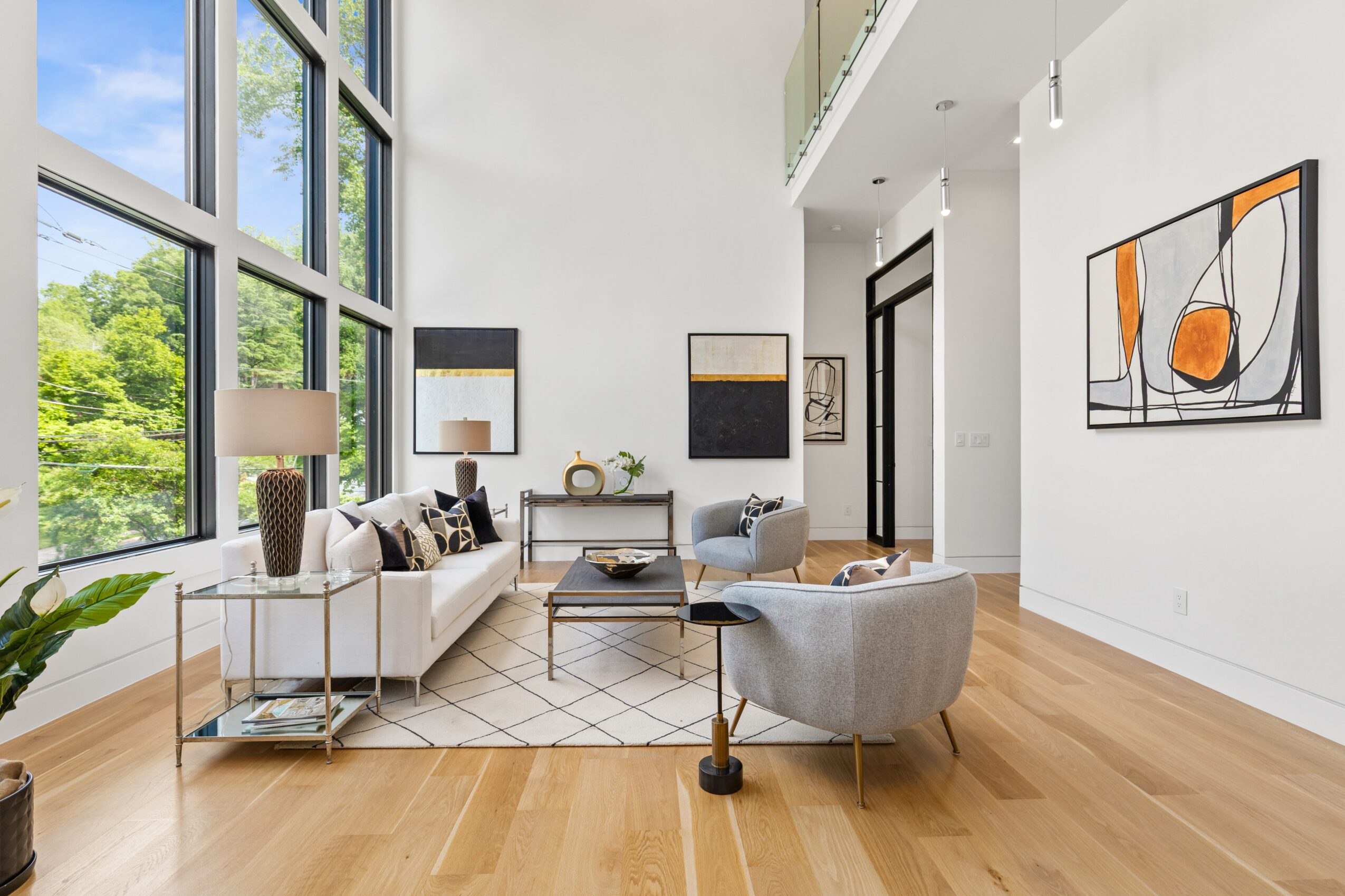 Living room with large windows, white sofa, armchair, coffee table, and abstract wall art, with hardwood floors and natural light.