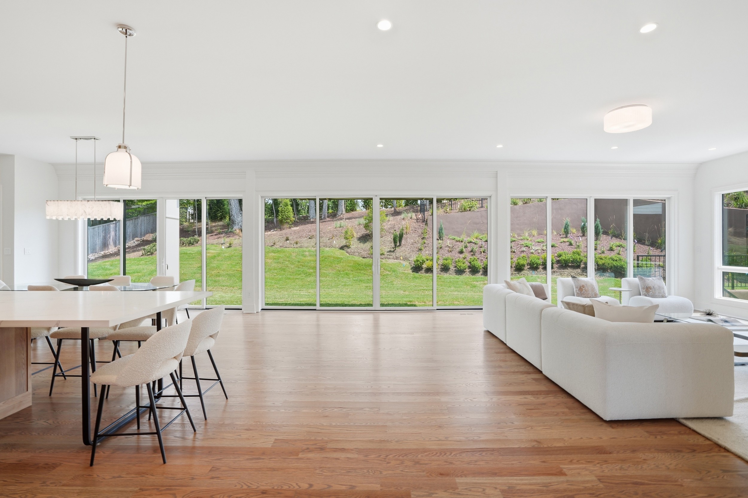 Spacious living room with large windows, white sofa, dining table, and wooden floor, overlooking a green outdoor area.