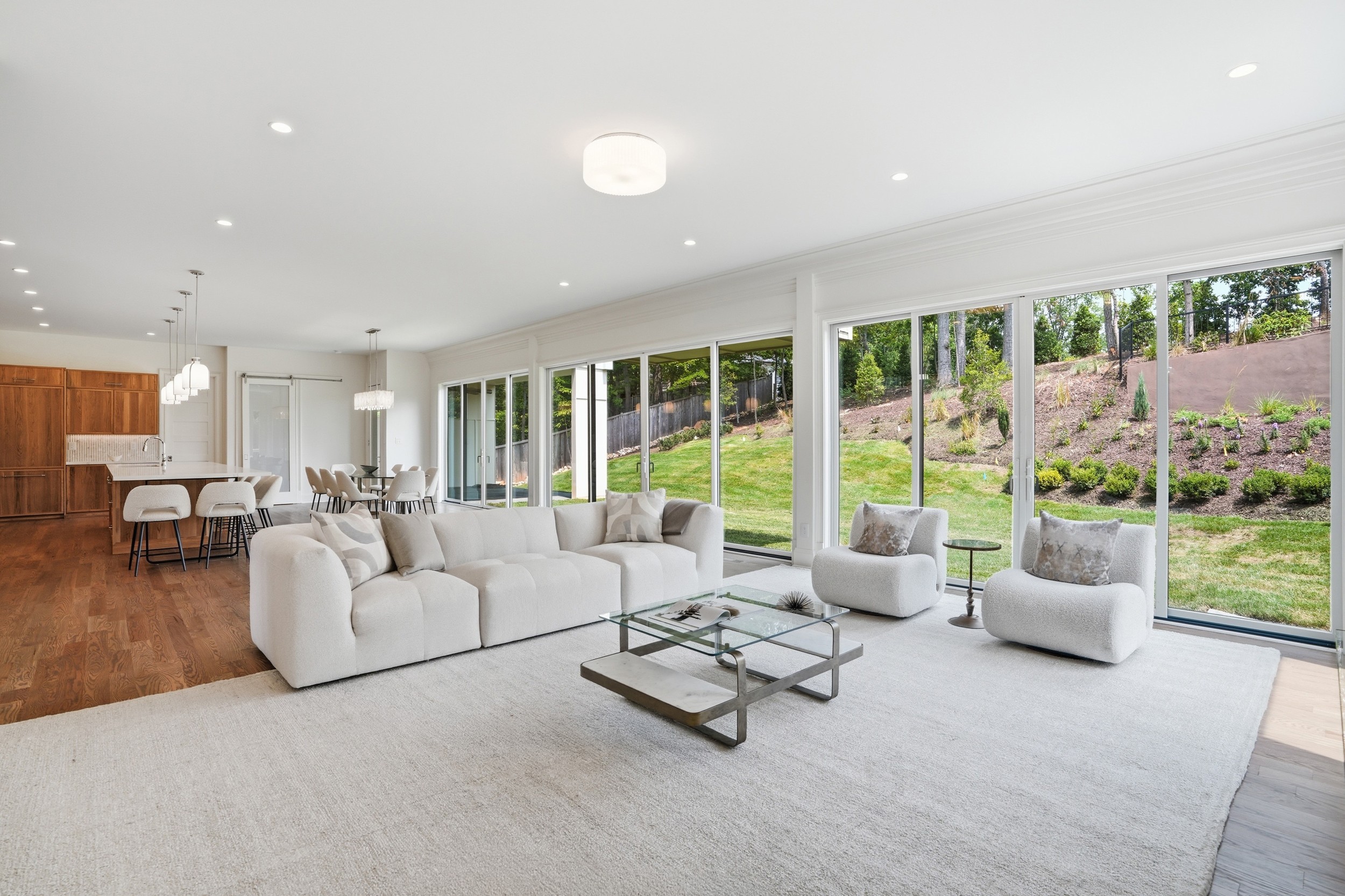 Spacious living room with large windows, white sofa, armchairs, glass coffee table, and view of garden outside.