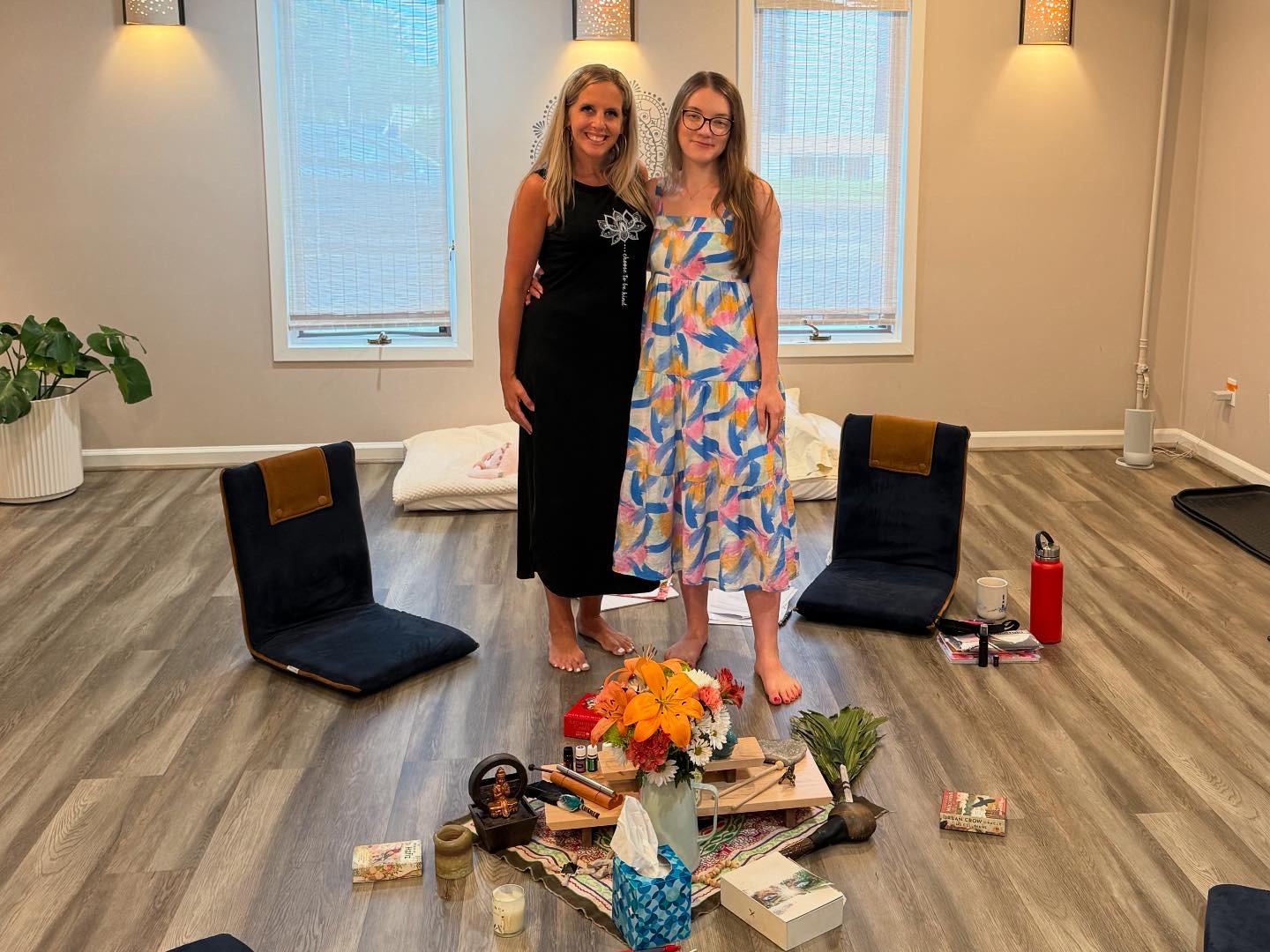 Two women stand in a room with wooden floors and two windows, surrounded by chairs, flowers, and various objects on the floor.