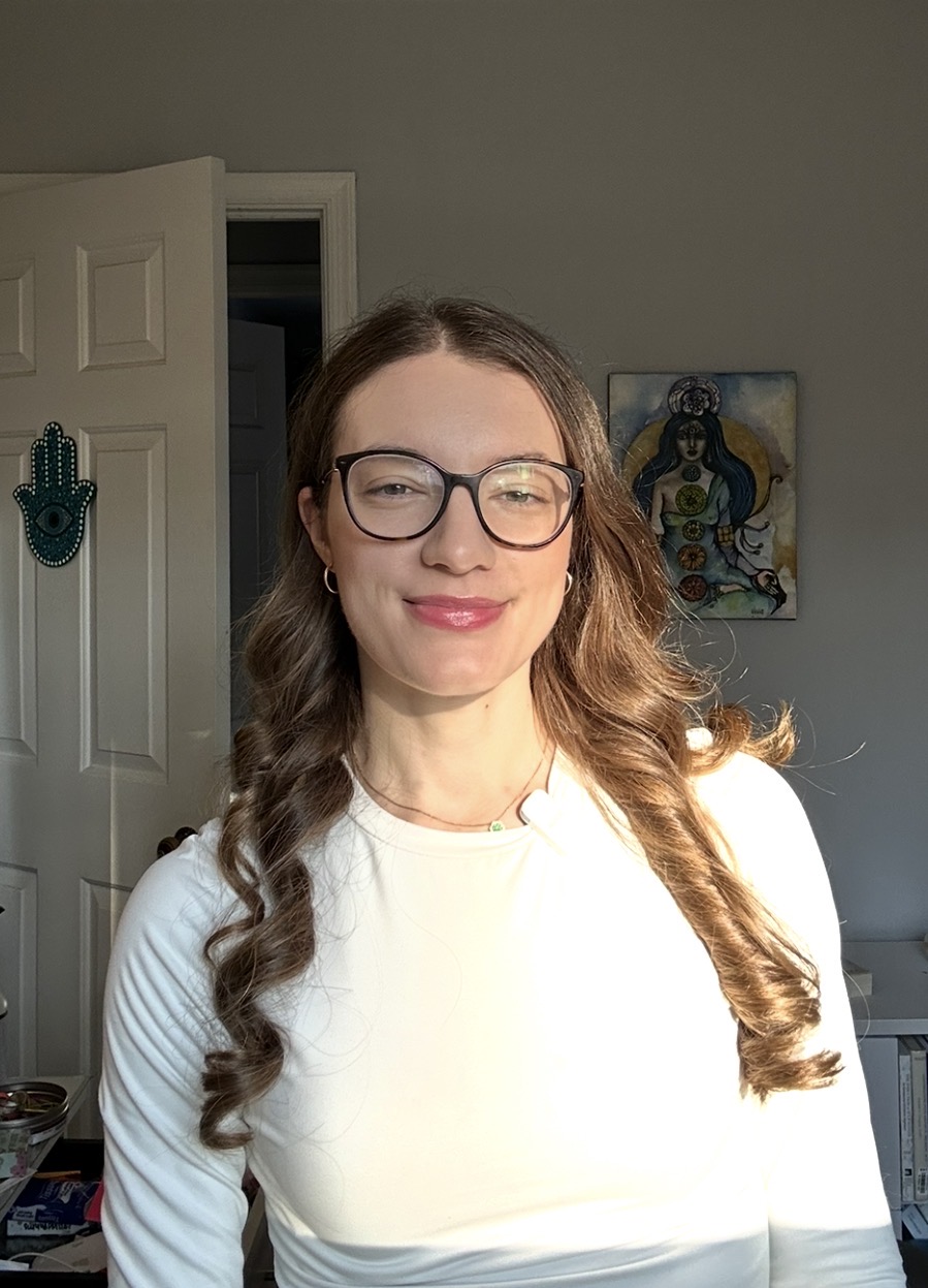 Young woman with glasses and wavy hair smiling in a room with a door and artwork on the wall.